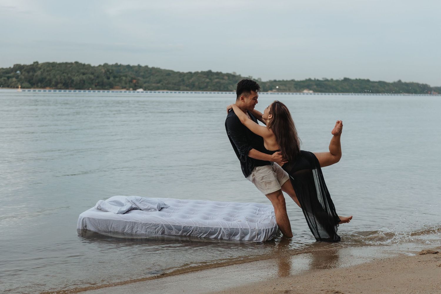 Couple poses romantically on mattress during seaside photoshoot.
