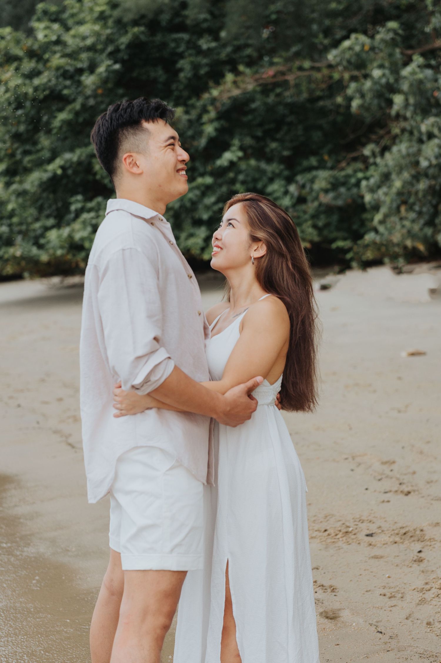 A couple in white outfits shares a joyful moment together on a beach.