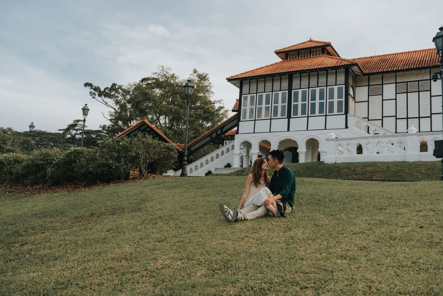 A couple sits together on a grassy hill in front of a large white building with terracotta roof tiles.