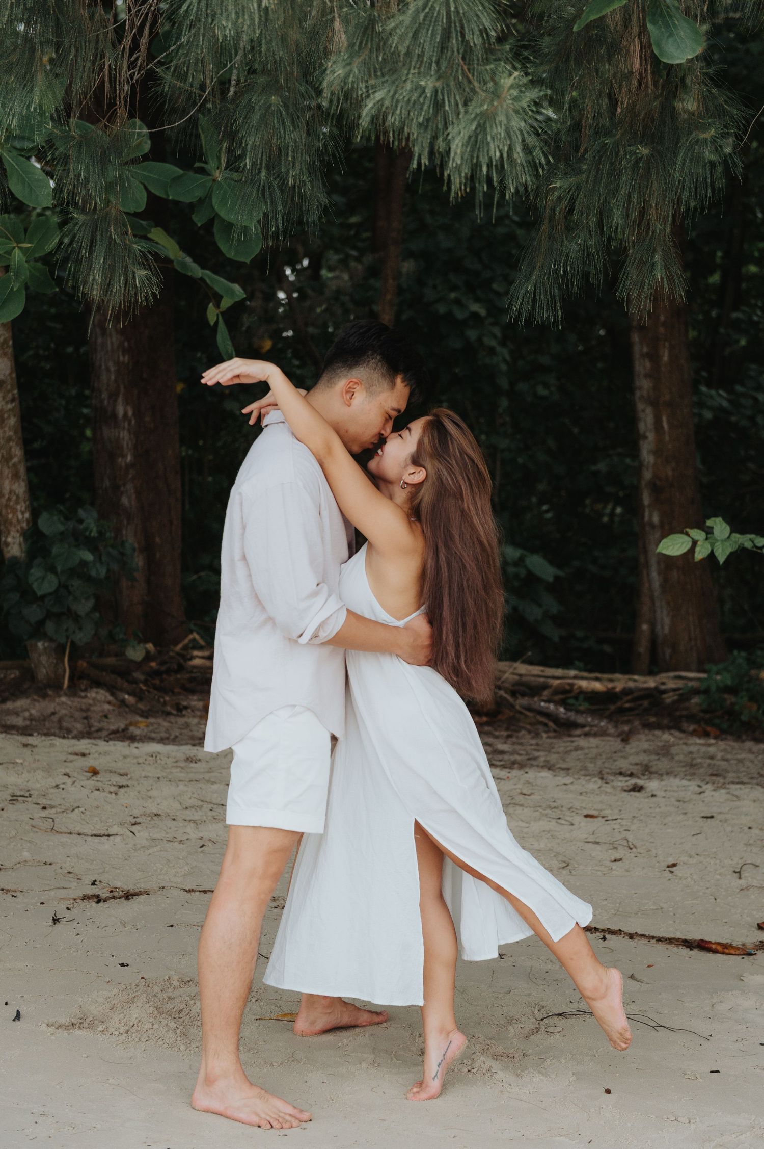 A couple in white outfits dances together on a tropical beach with pine trees in the background.