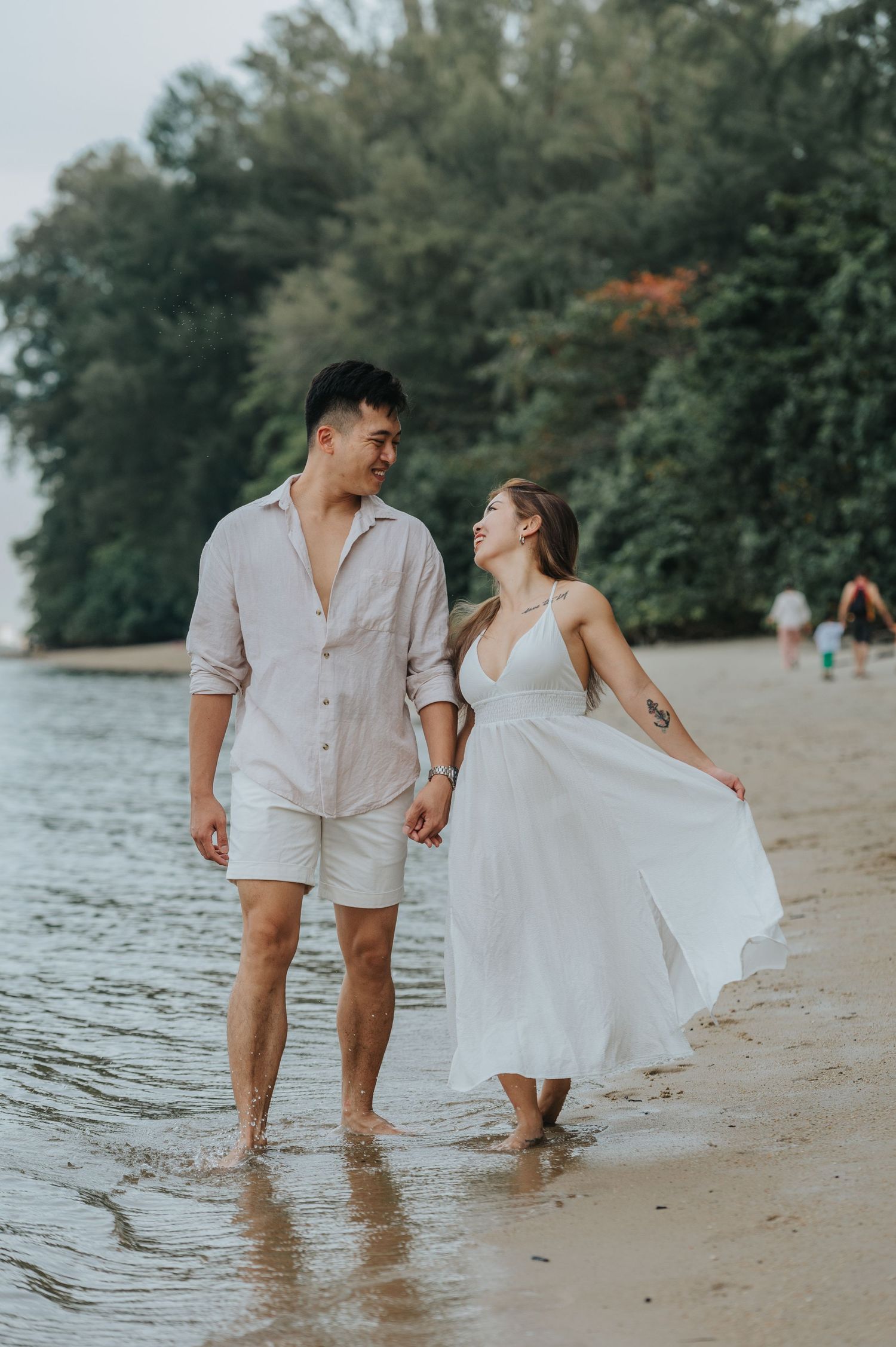 A couple in matching white outfits walks along a beach shoreline with lush greenery in the background.