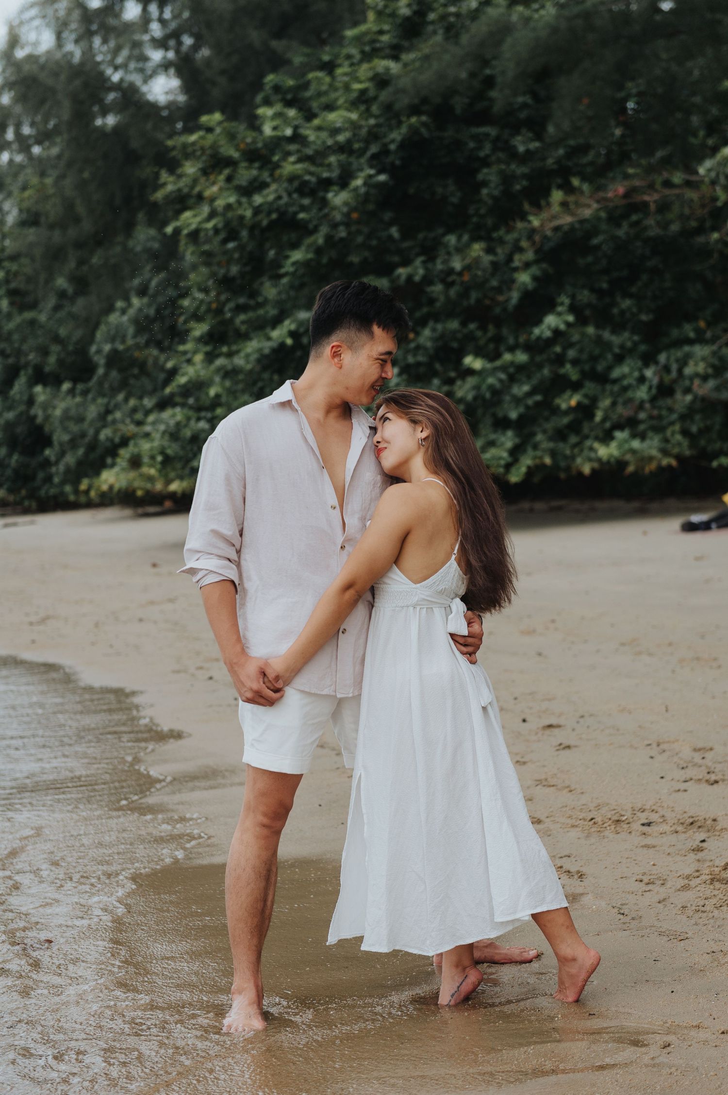 A couple in white clothing shares an intimate moment while standing barefoot on a beach.