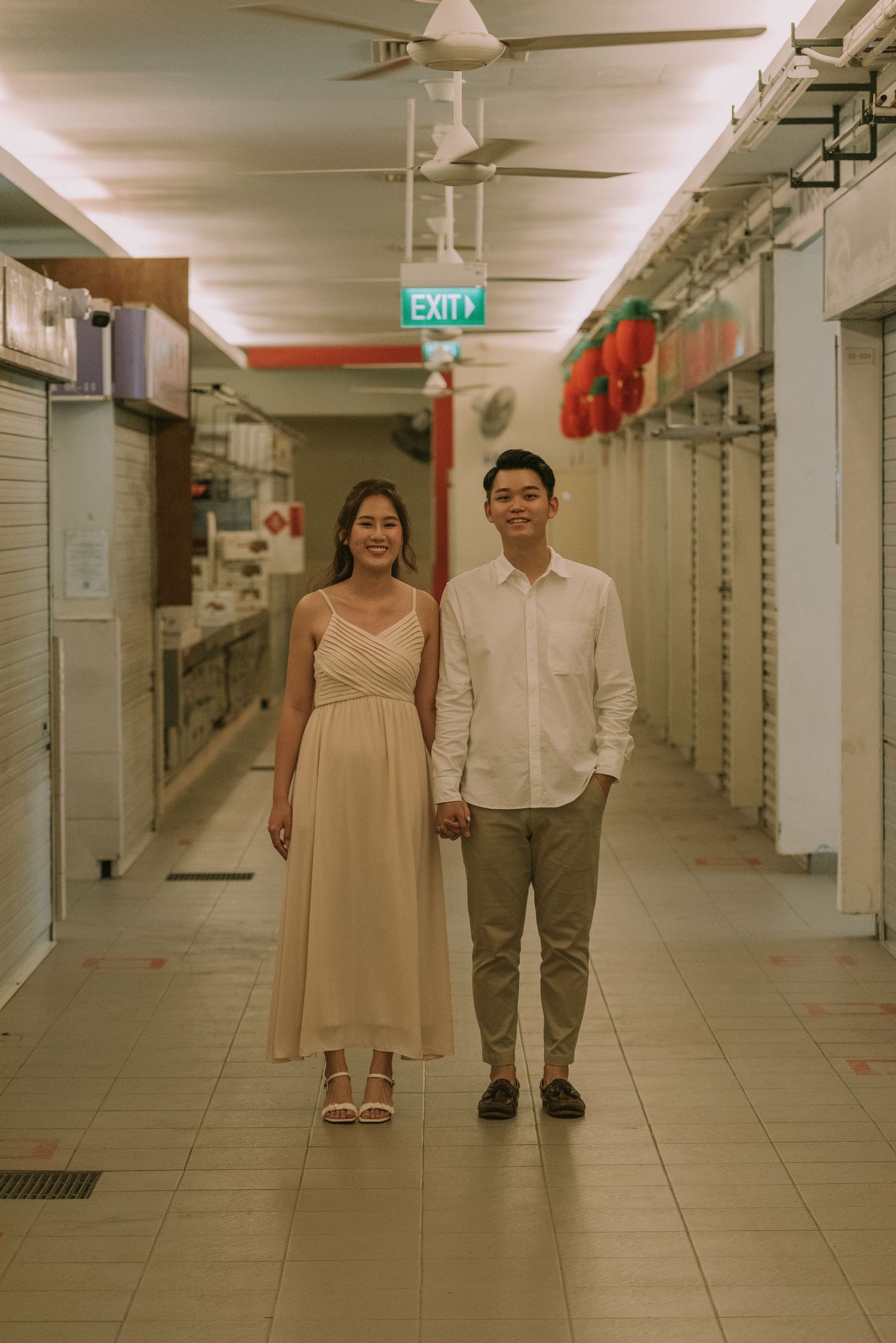 Couple in cream and white outfits posing in a brightly lit corridor with an exit sign.