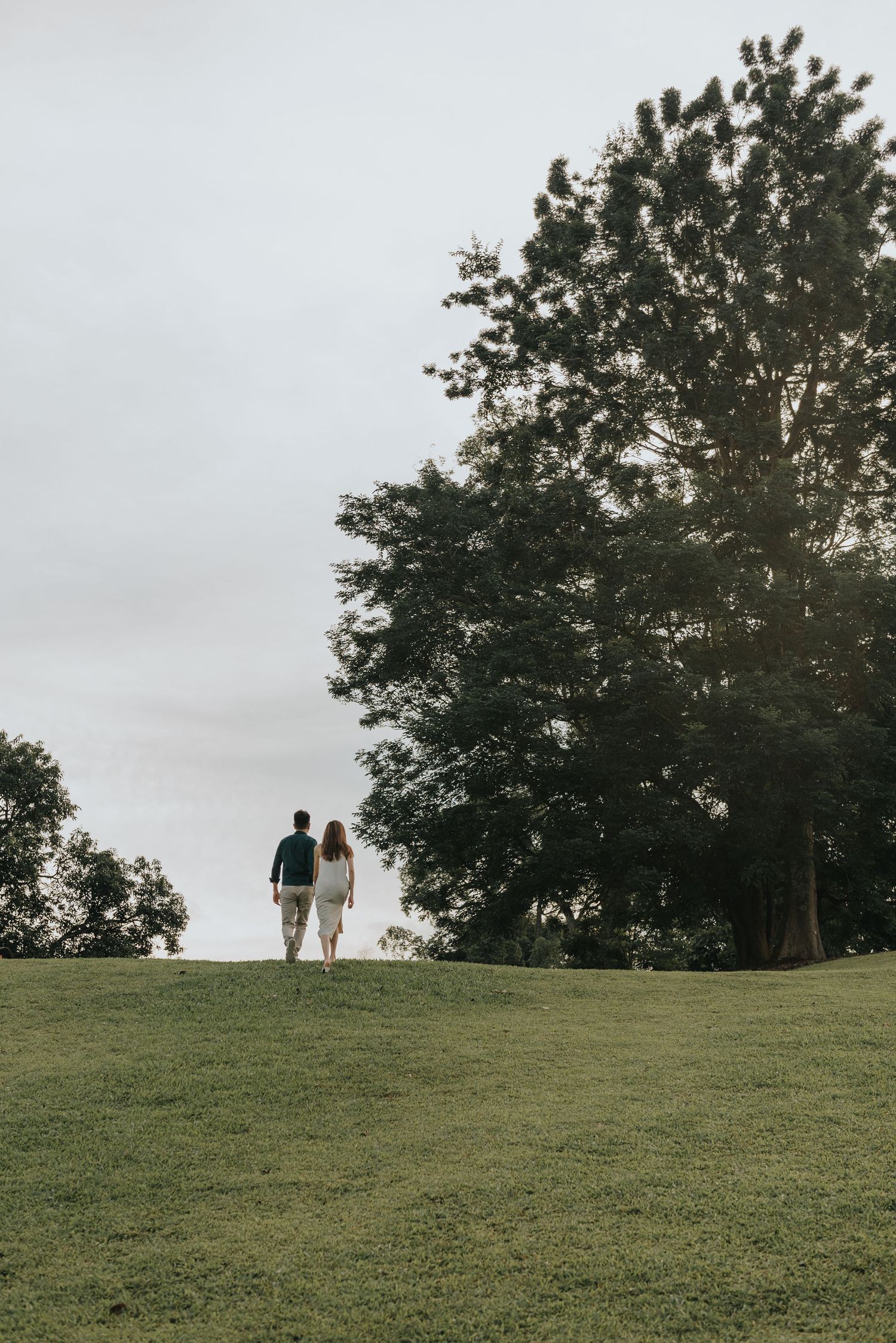 Two people walk up a grassy slope with large trees in the background against an overcast sky.