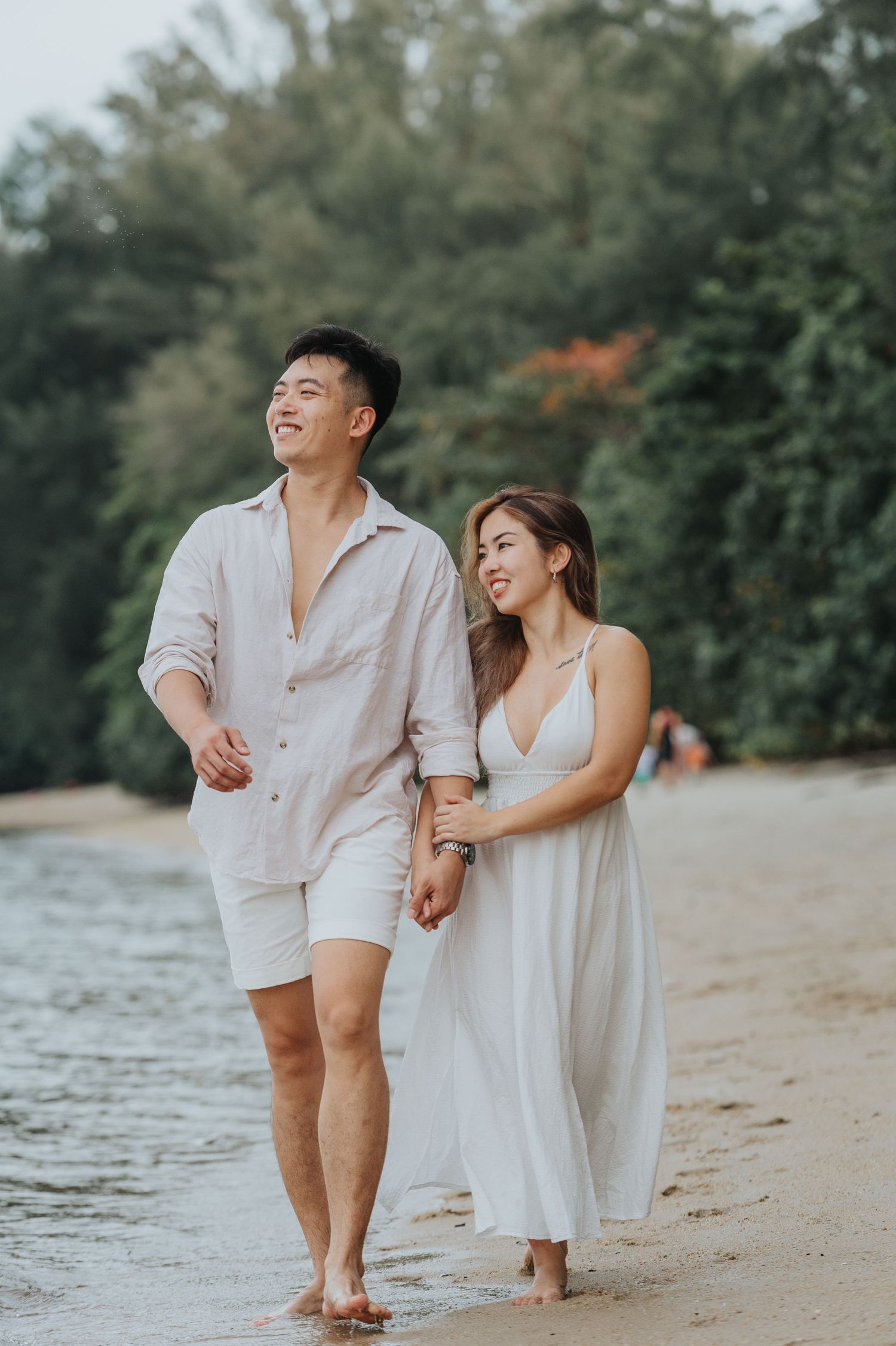 A happy couple walks hand in hand on the beach wearing coordinated white outfits.