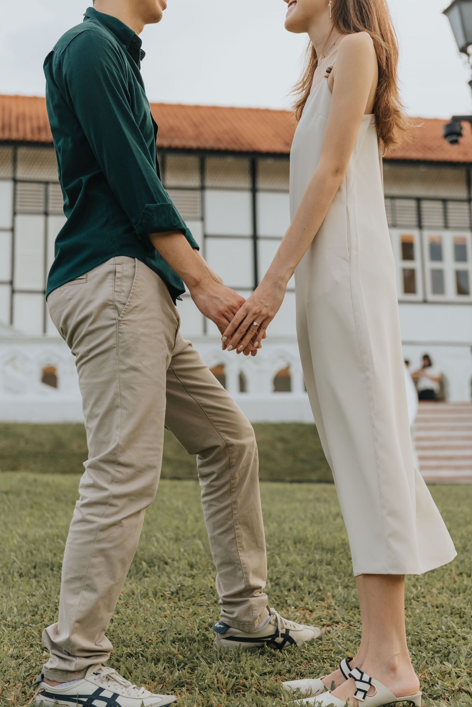 Two people in casual outfits standing close together on grass near colonial architecture.