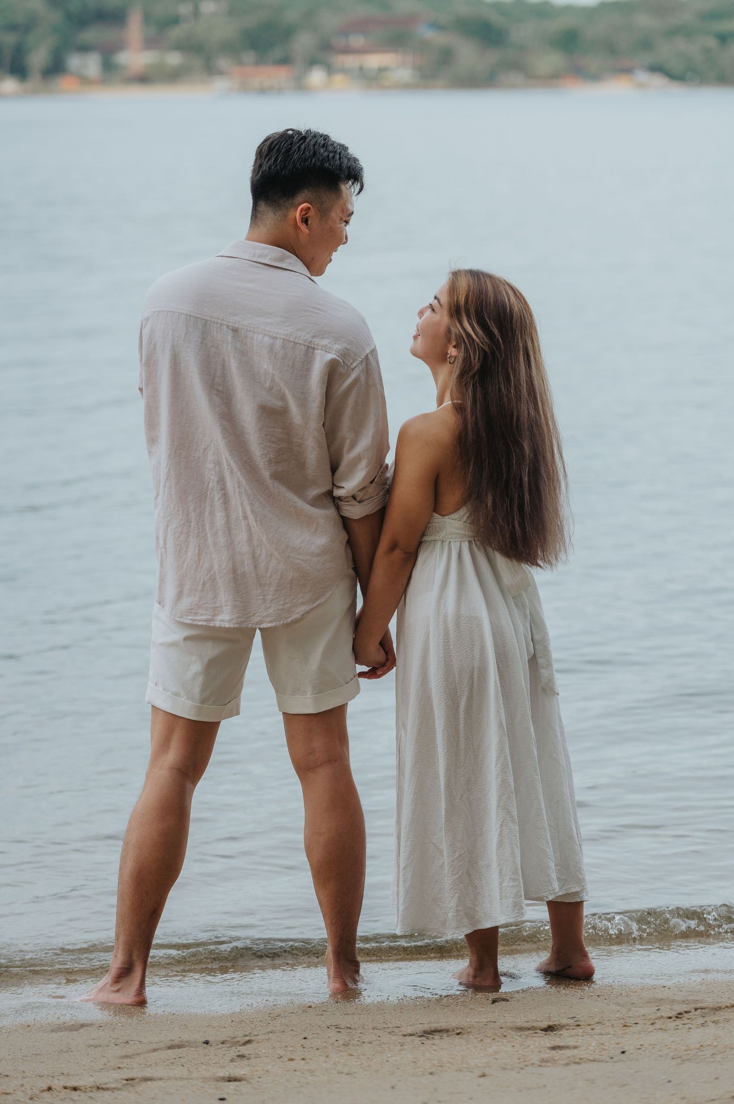 A romantic couple stands barefoot on a beach shore gazing at each other in light colored summer outfits.
