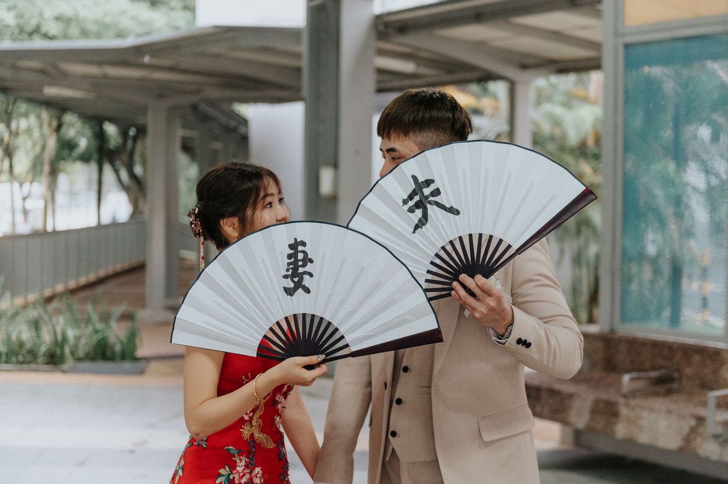 Couple holding traditional Chinese fans during wedding photoshoot under covered walkway.