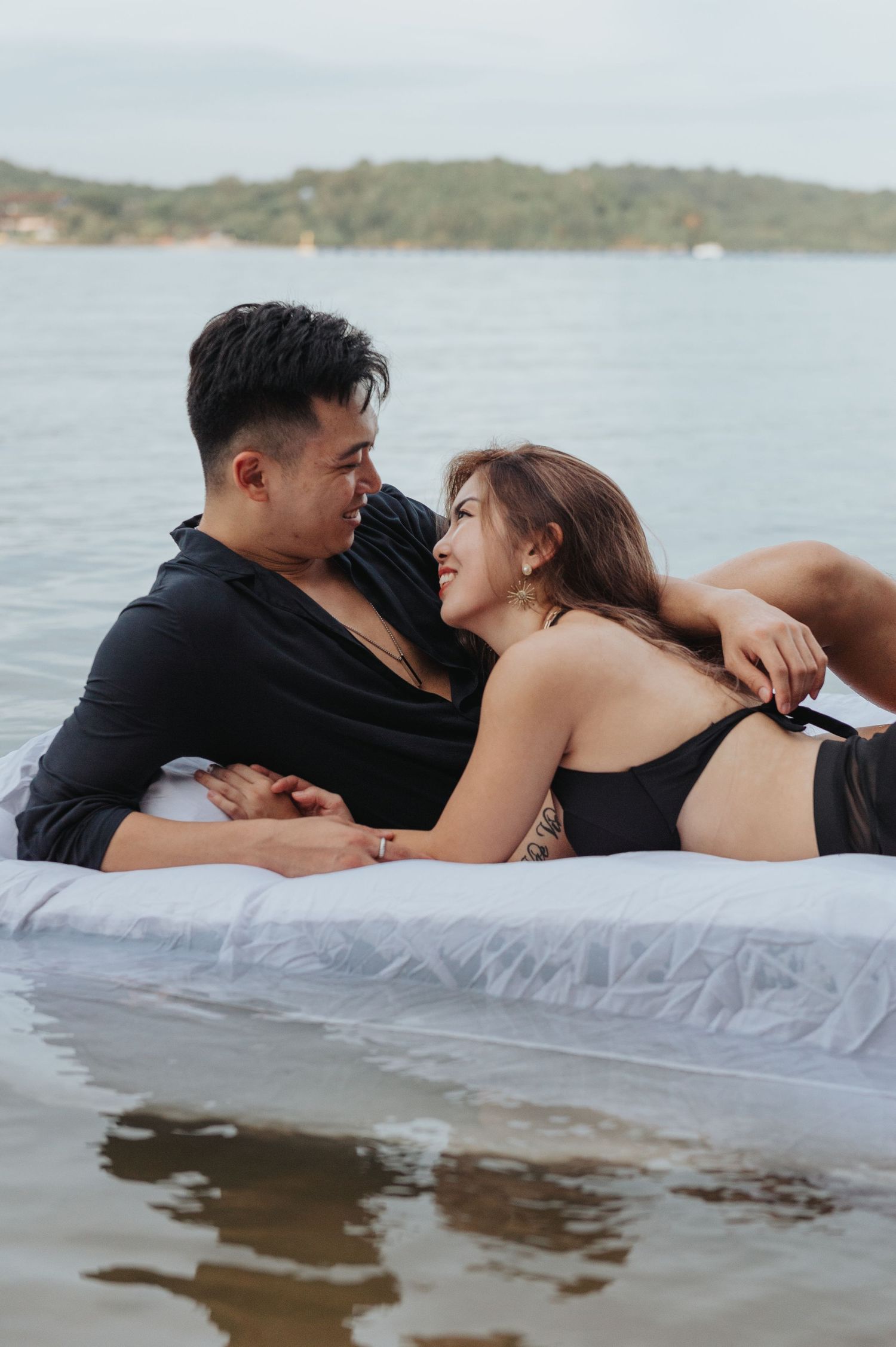 A couple relaxes intimately on a white mattress at the water's edge.