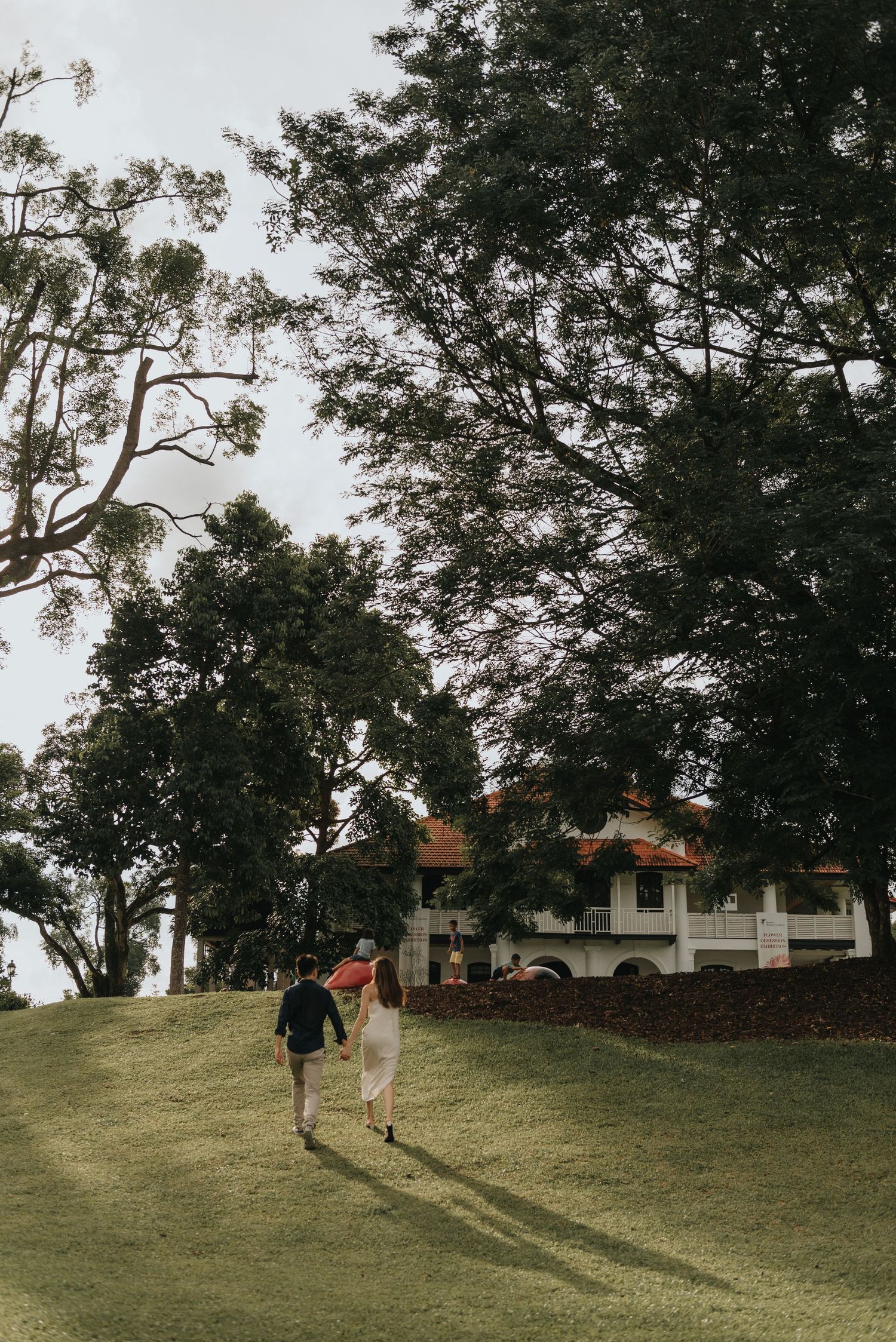 A couple ascends a grassy hill towards a white colonial mansion under moody skies.