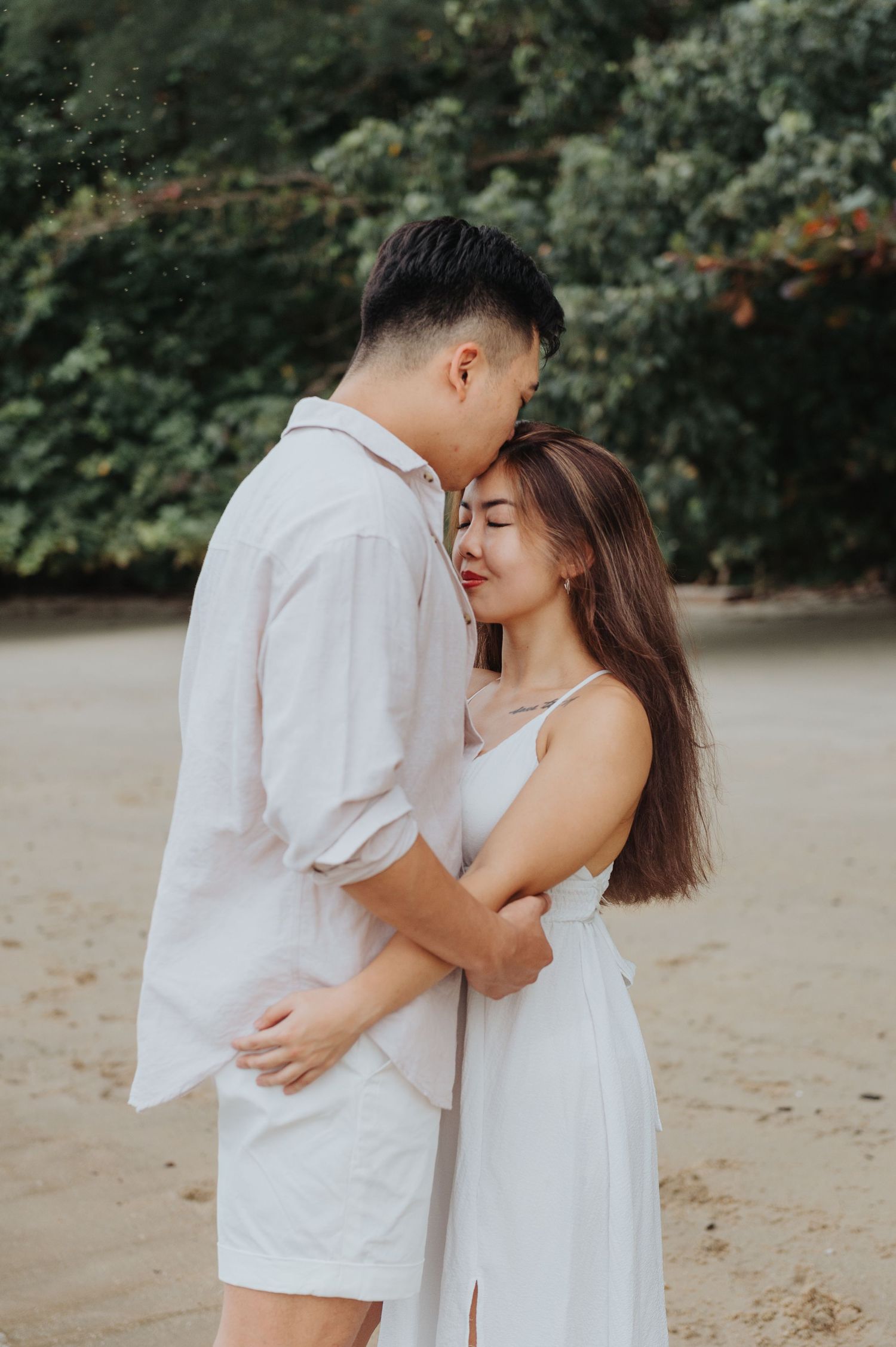 A romantic couple in light colored outfits embraces on a sandy beach with lush foliage behind them.