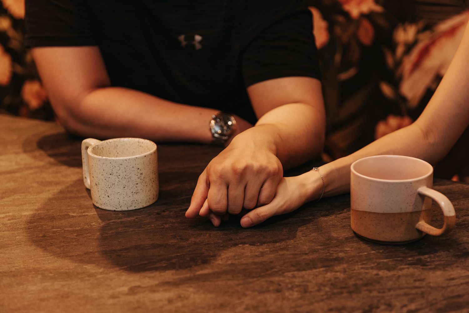 Two coffee cups sit on a wooden table as hands clasp together in an intimate moment.