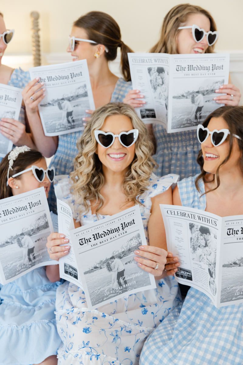 Bride in heart-shaped sunglasses reading custom wedding newspaper with bridesmaids at Jekyll Island Club. Fun, candid moment.