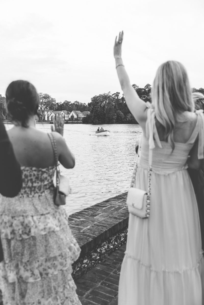 Wedding guests wave as bride and groom arrive by boat at Frederica Golf Club Boathouse on St. Simons Island, captured by photographer.