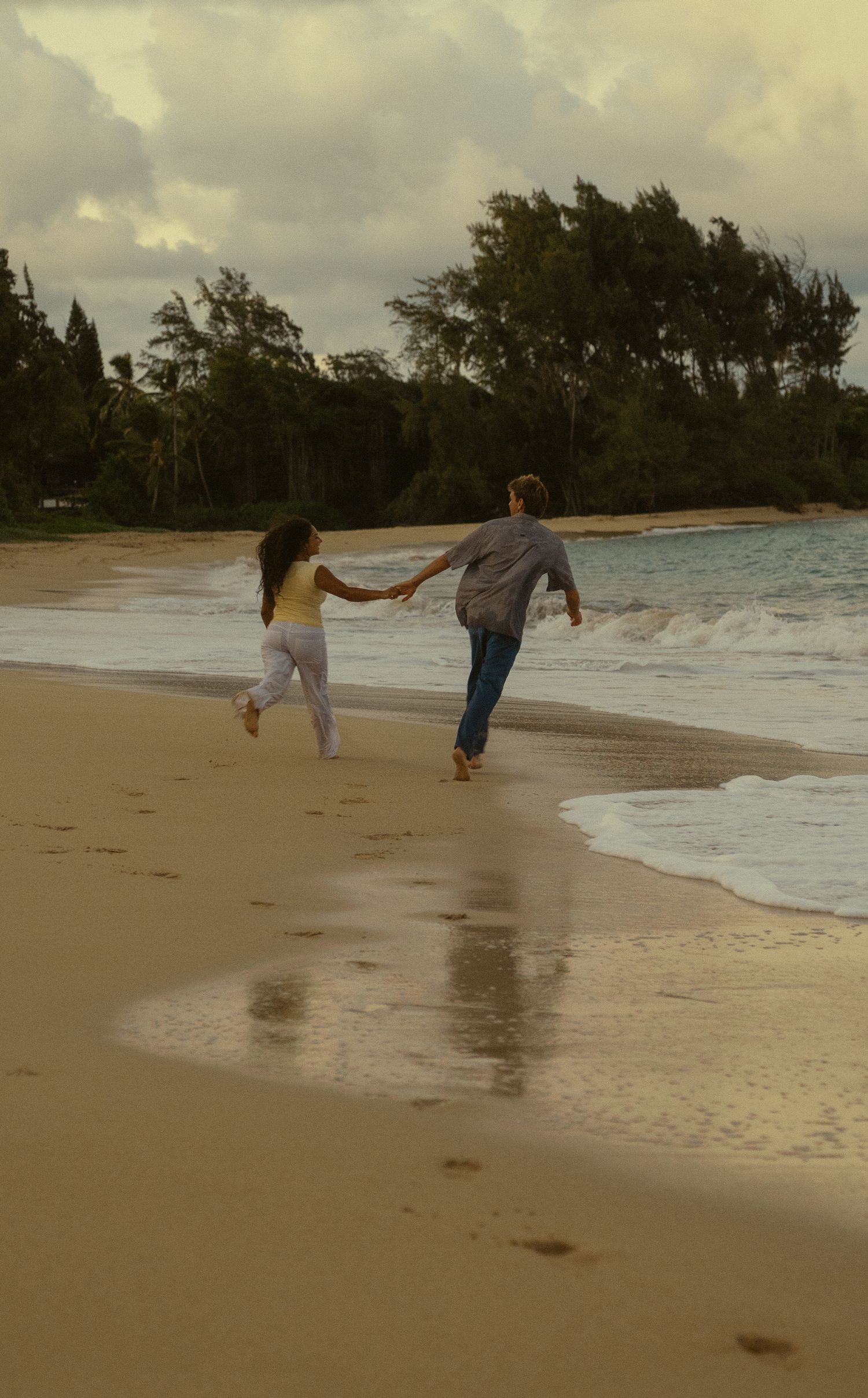 A playful couple runs along the shoreline as waves crash on a tropical beach.