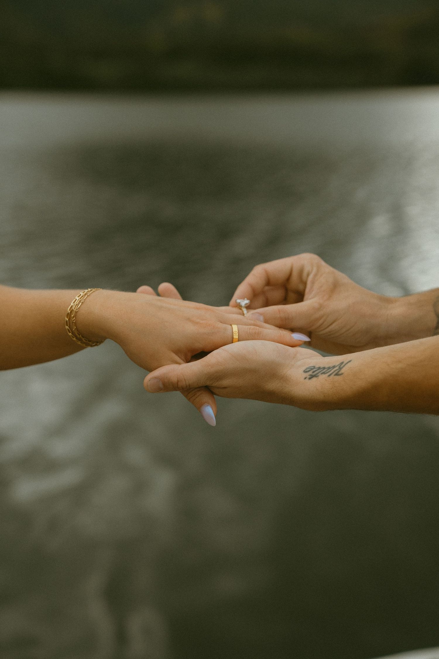 Two hands reaching towards each other with jewelry against a blurred natural background.