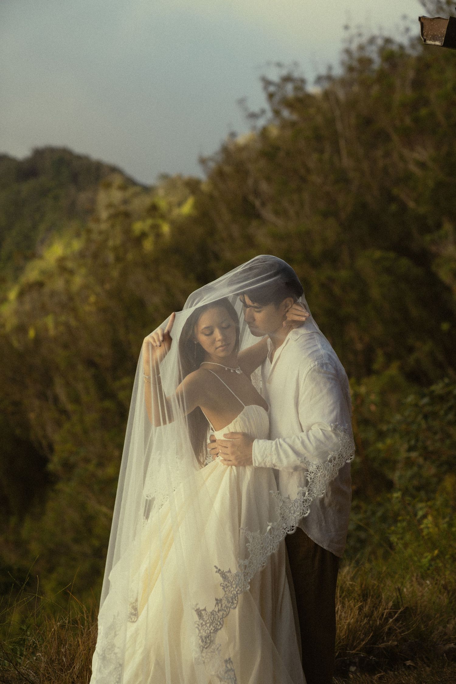A romantic wedding couple embraces outdoors during golden hour with a flowing lace veil and natural backdrop.