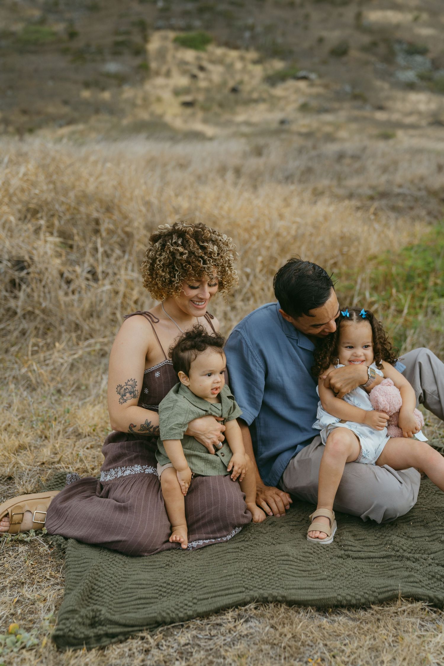 Family relaxes together on a blanket in a natural outdoor setting with mountains in background.