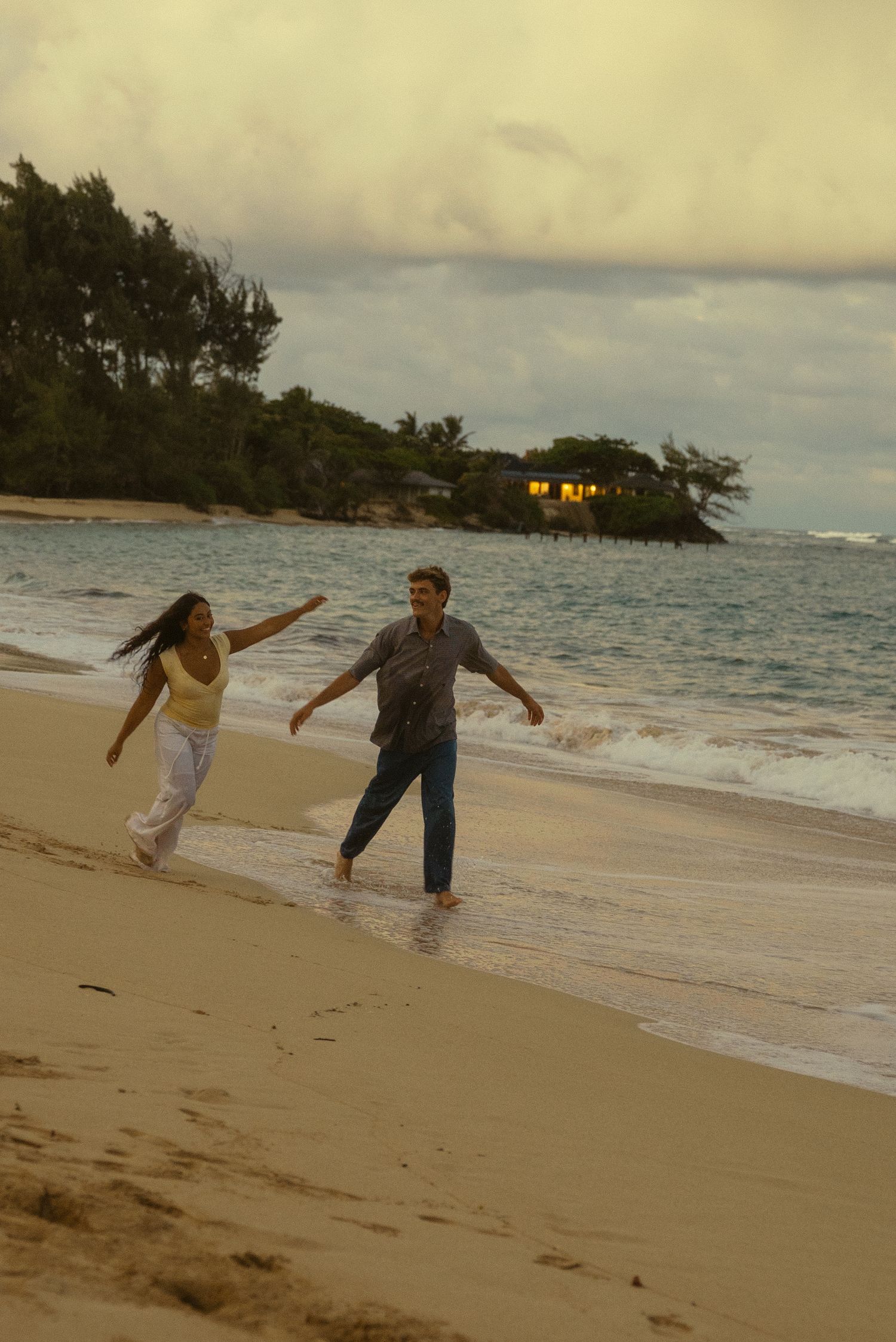 Two people dance and play in the shallow ocean waves on a tropical beach at sunset.