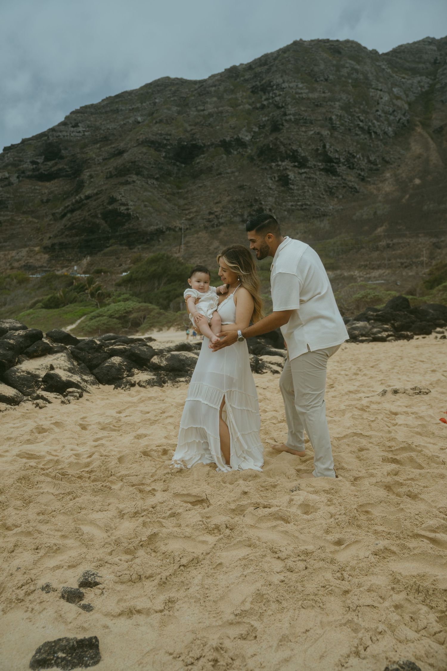 Candid family moments captured on a sandy beach with rocky cliffs in Hawaii.