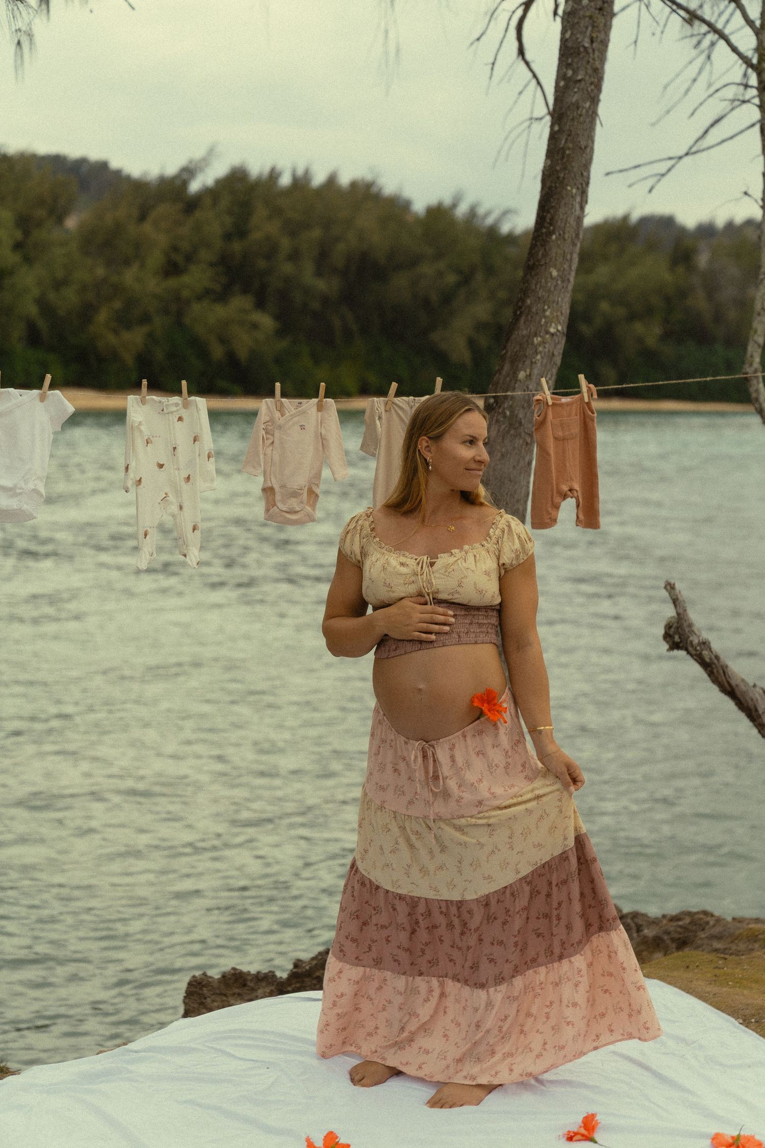 A person in a flowy dress sits on a white blanket by the ocean with clotheslines in view.