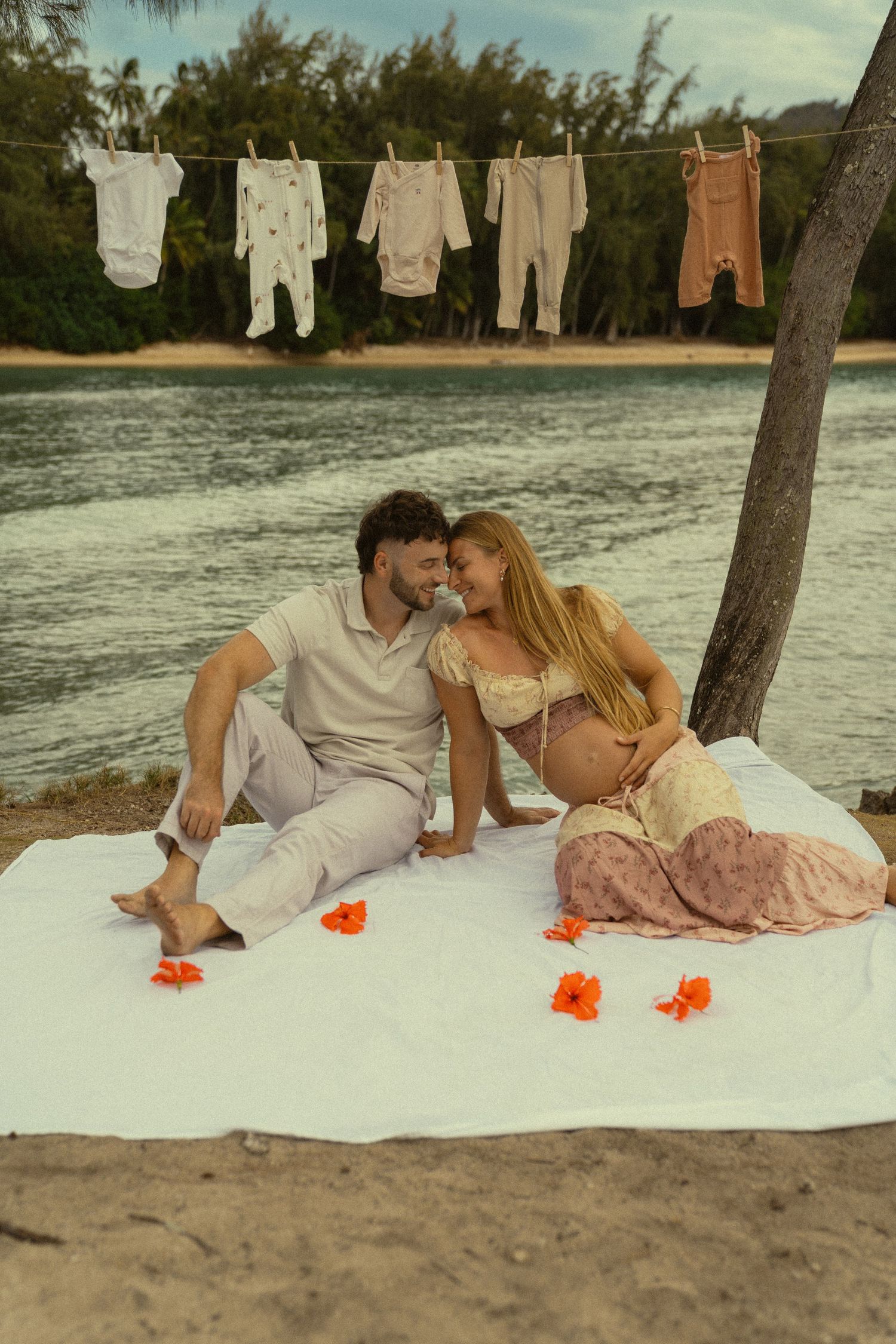 A couple relaxes on a white blanket with orange flowers and vintage clothes hanging nearby.