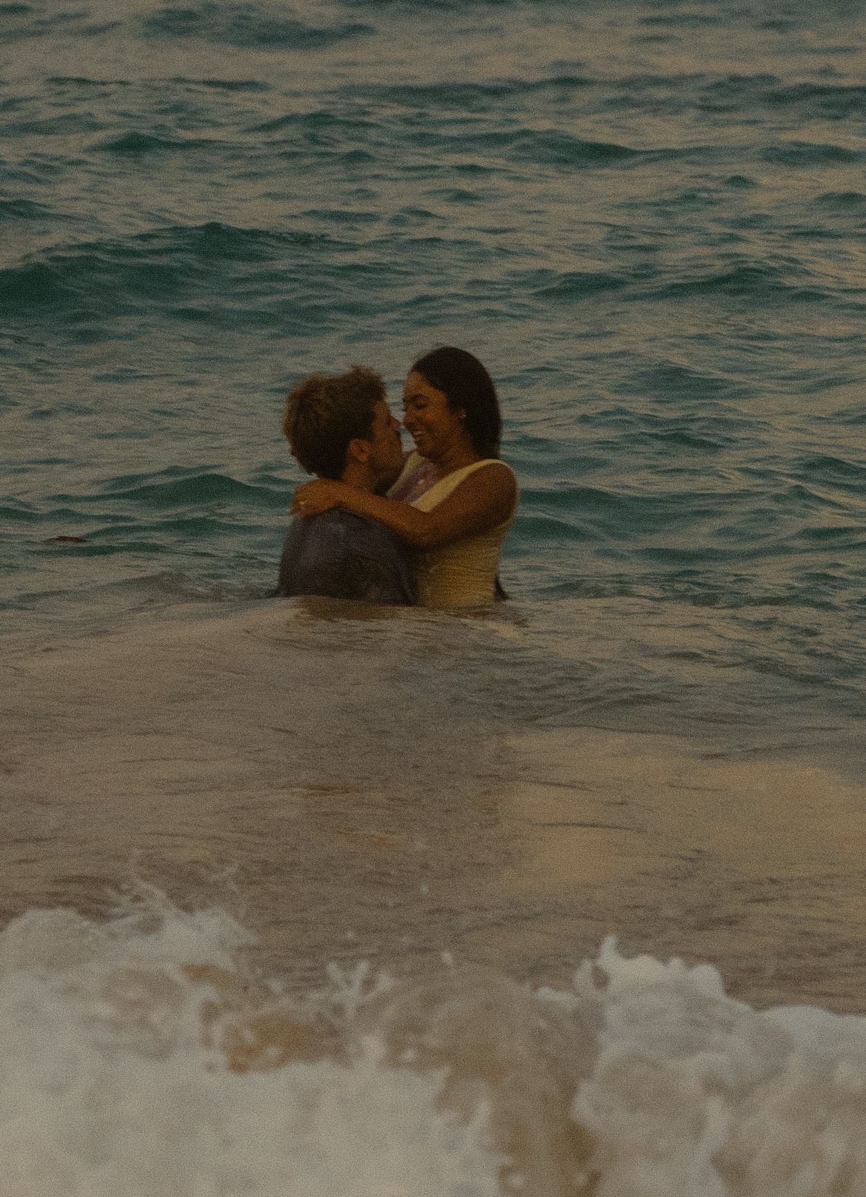 A romantic couple embraces in the ocean waves at sunset.