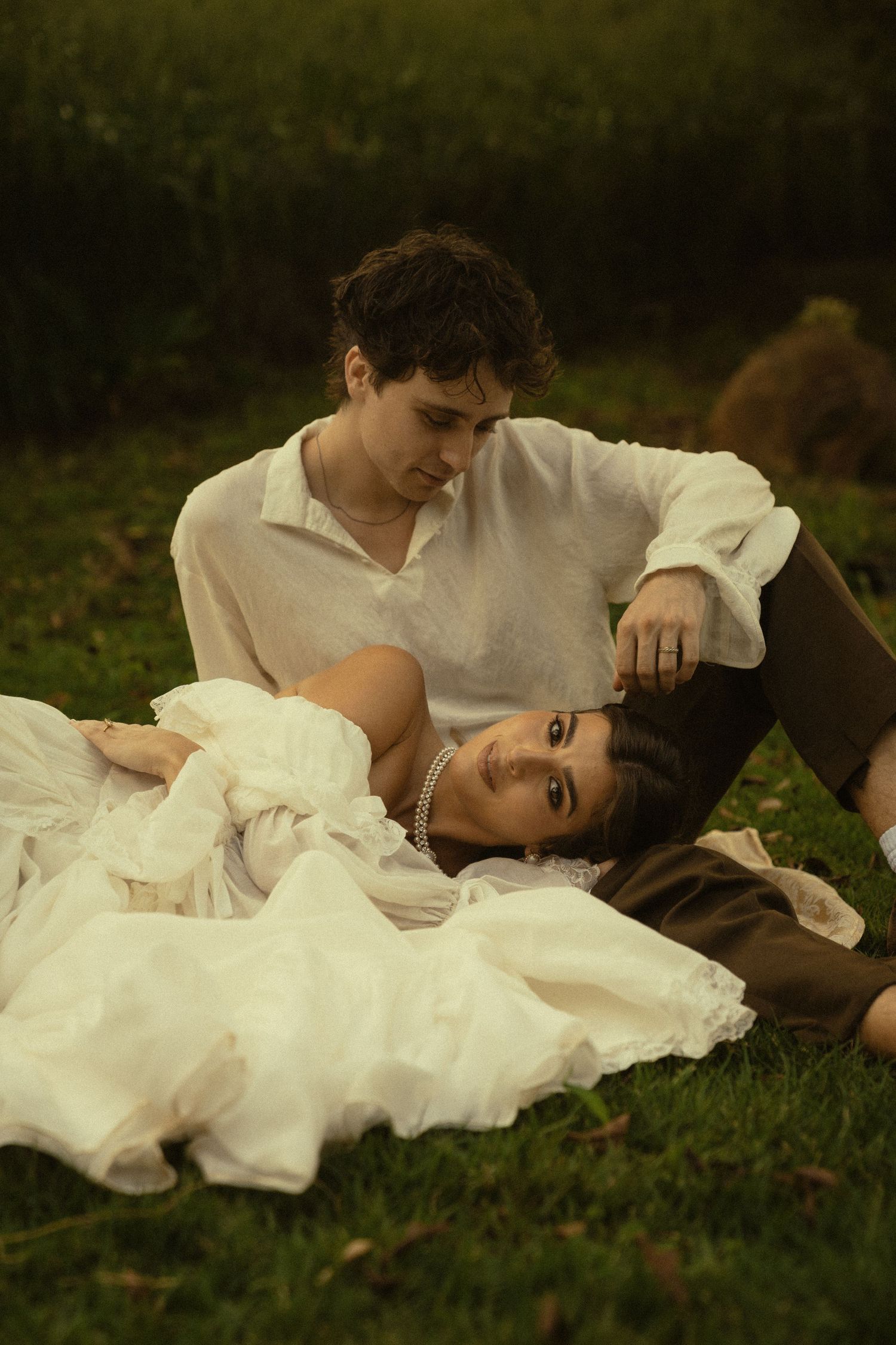 A romantic couple lying in grass wearing vintage-style white clothing in a dreamy outdoor setting.