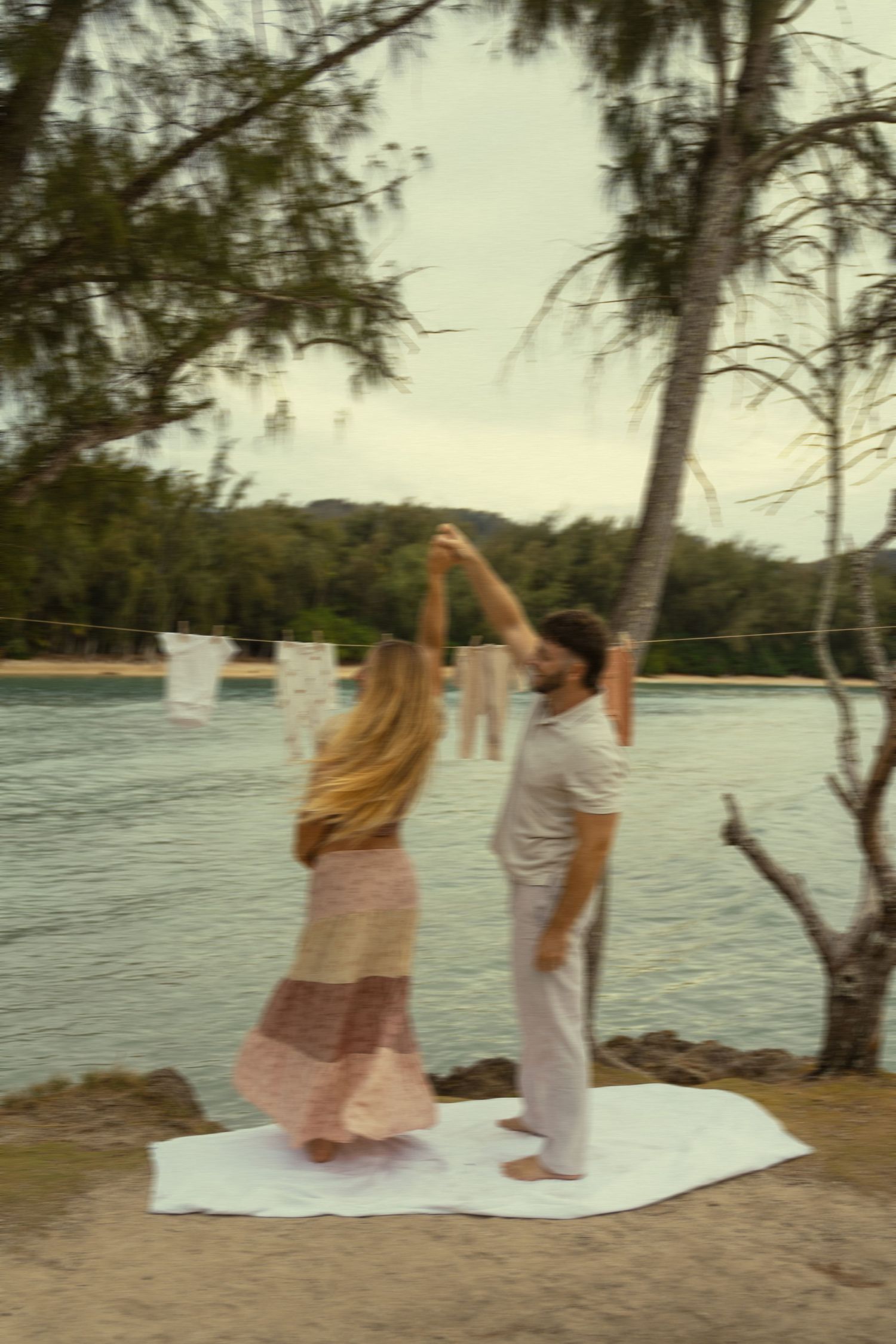 Two people dance together on a beach blanket with the ocean and trees behind them.
