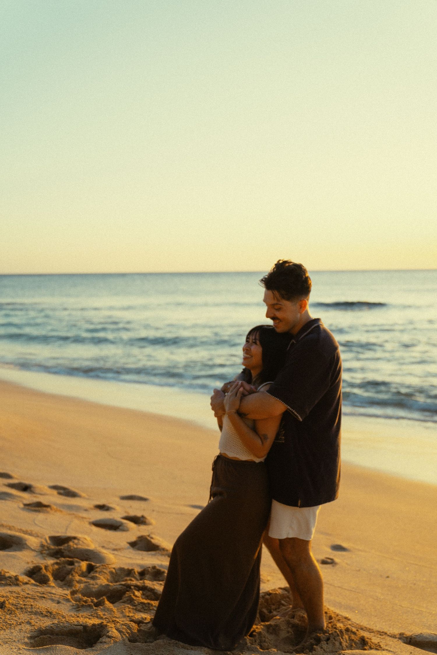 A couple embraces on a serene beach during sunset with gentle waves in the background.