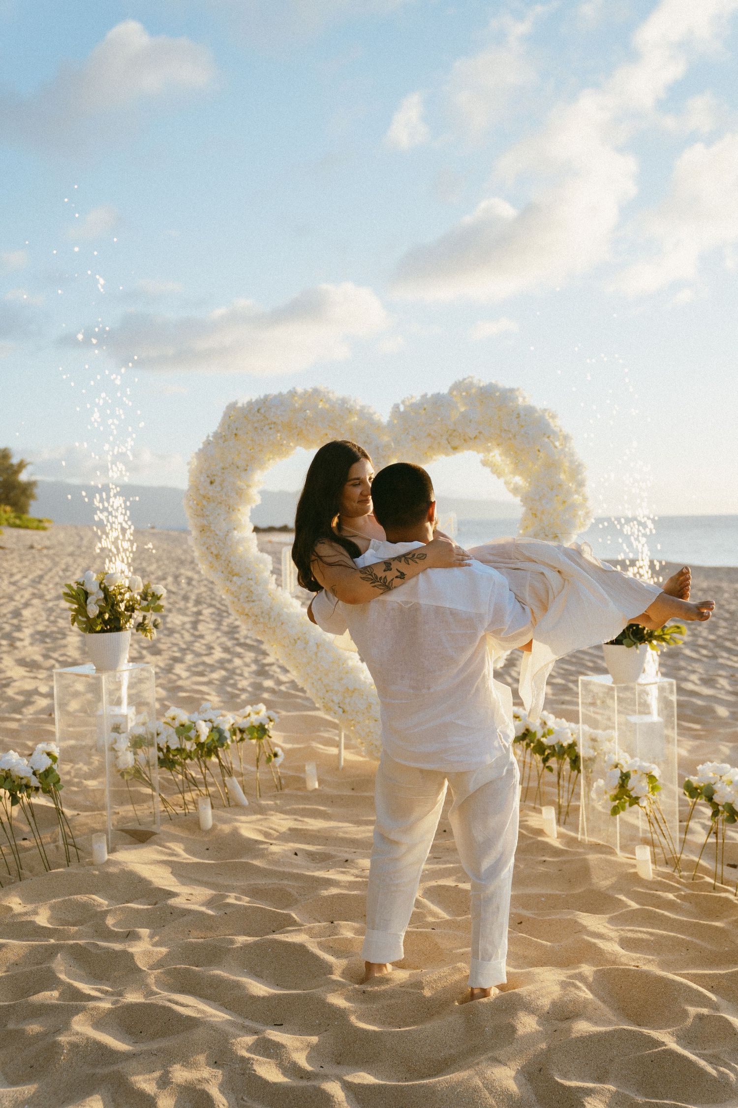 Two figures in white dance joyfully on a beach surrounded by floral decorations.