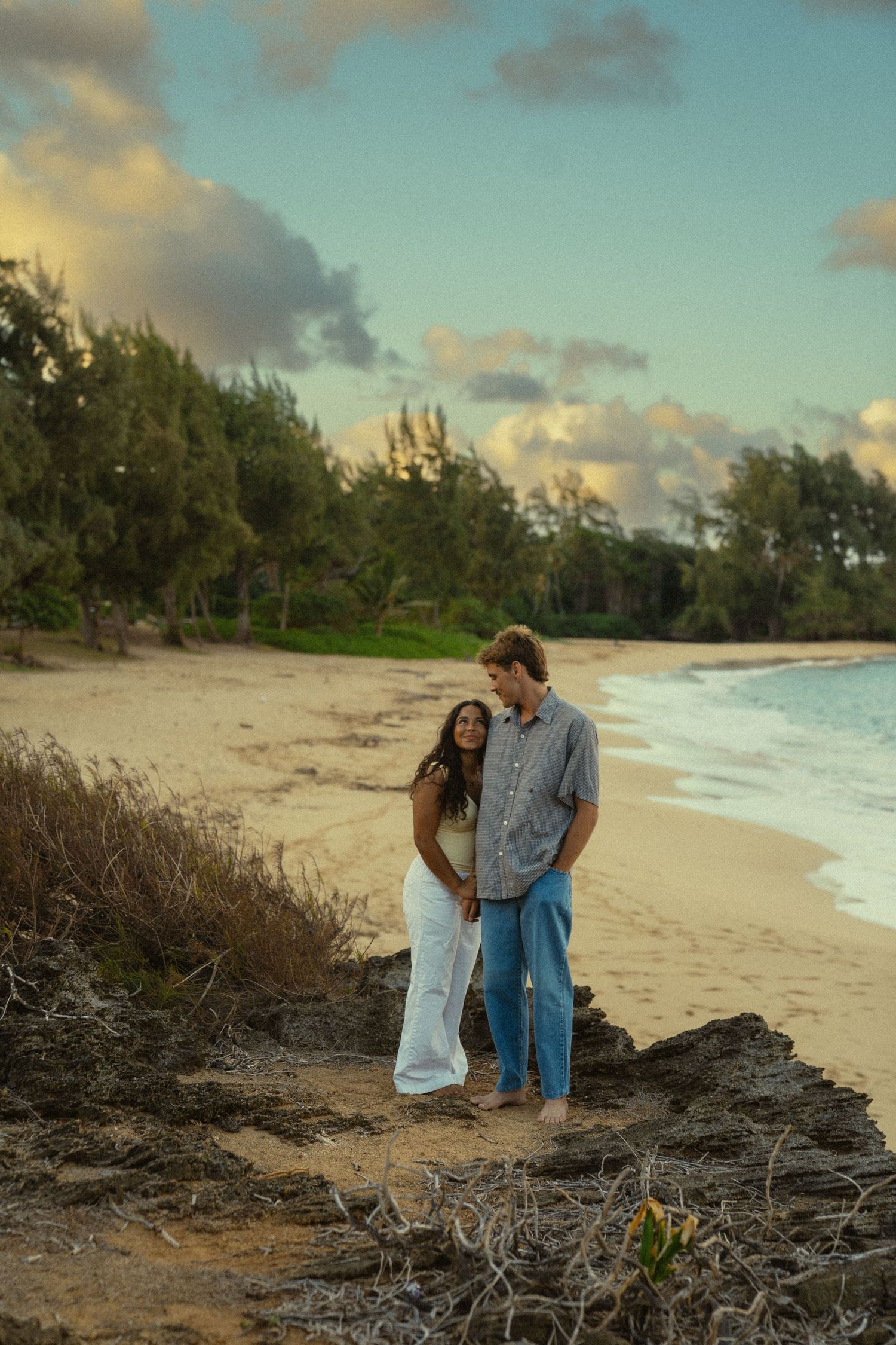A romantic couple shares an intimate moment on a scenic beach at sunset with palm trees in the background.
