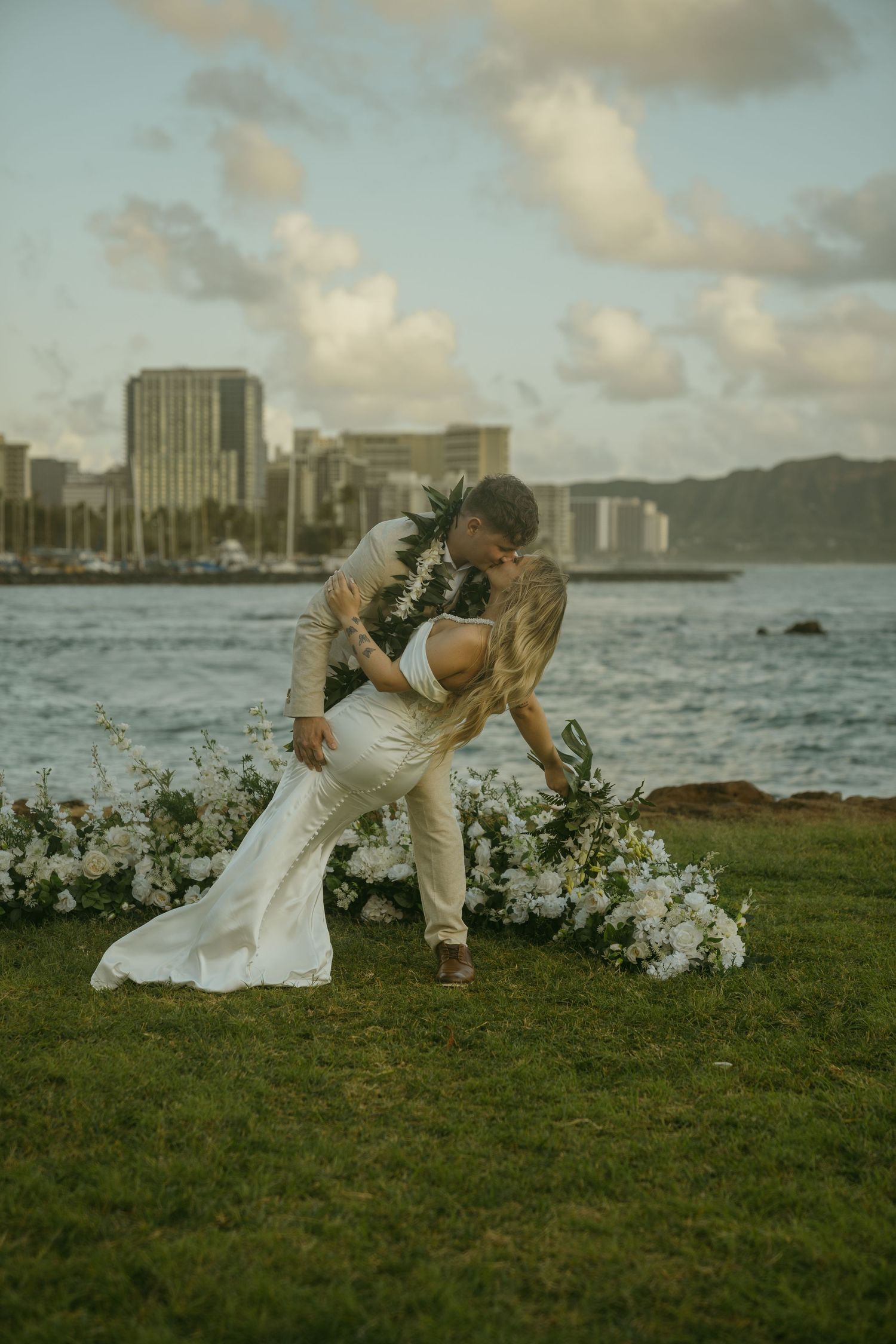 Couple in wedding attire dancing on grass with urban waterfront backdrop.