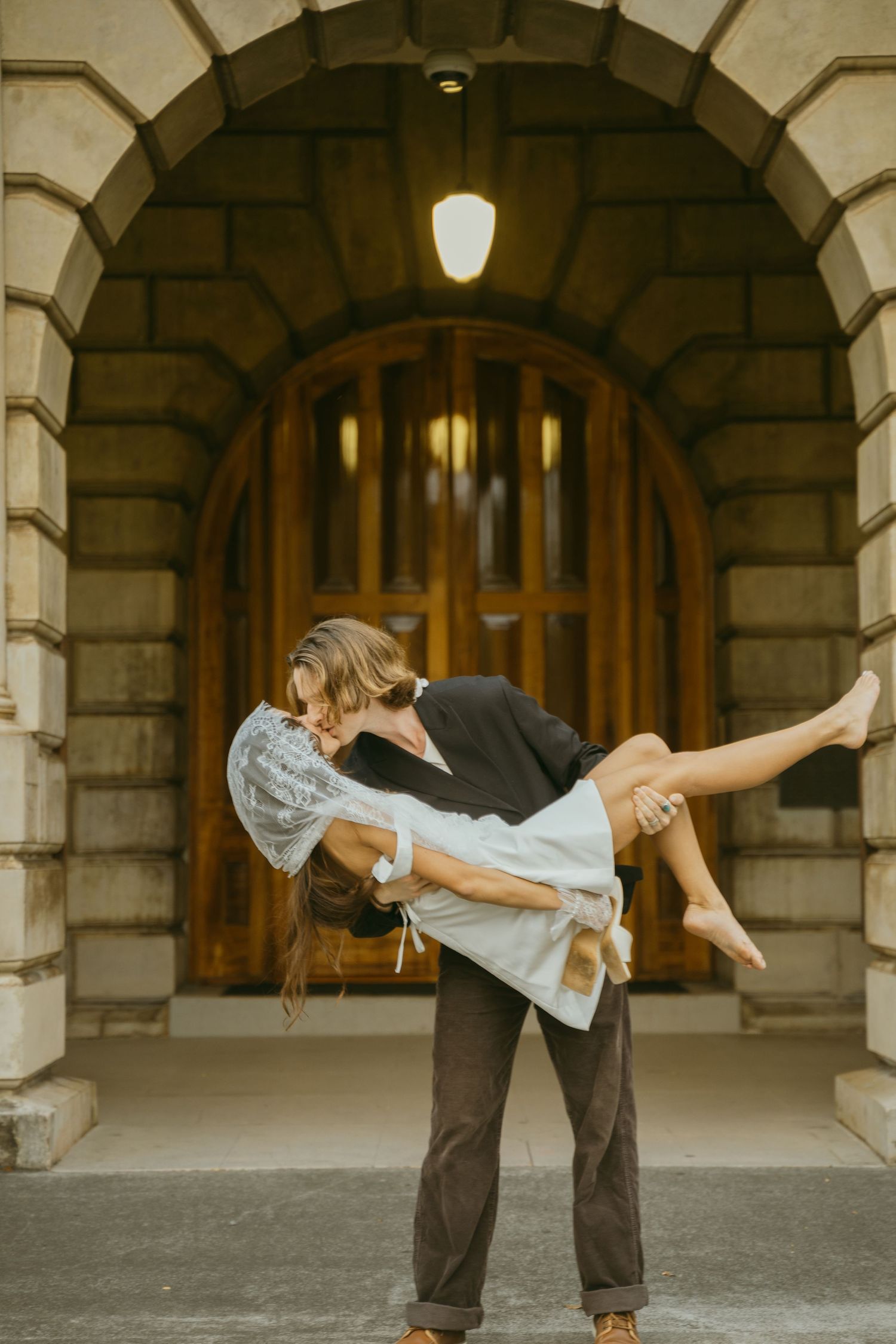 Playful wedding couple performing a dramatic dip pose under a stone archway.