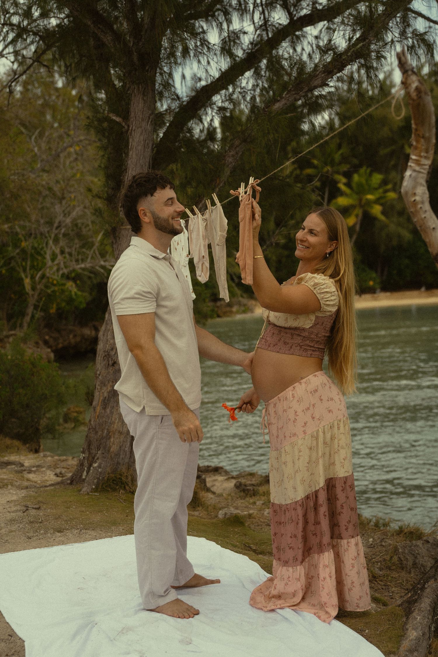 A couple dances together on a white blanket by the water's edge during a romantic outdoor photoshoot.