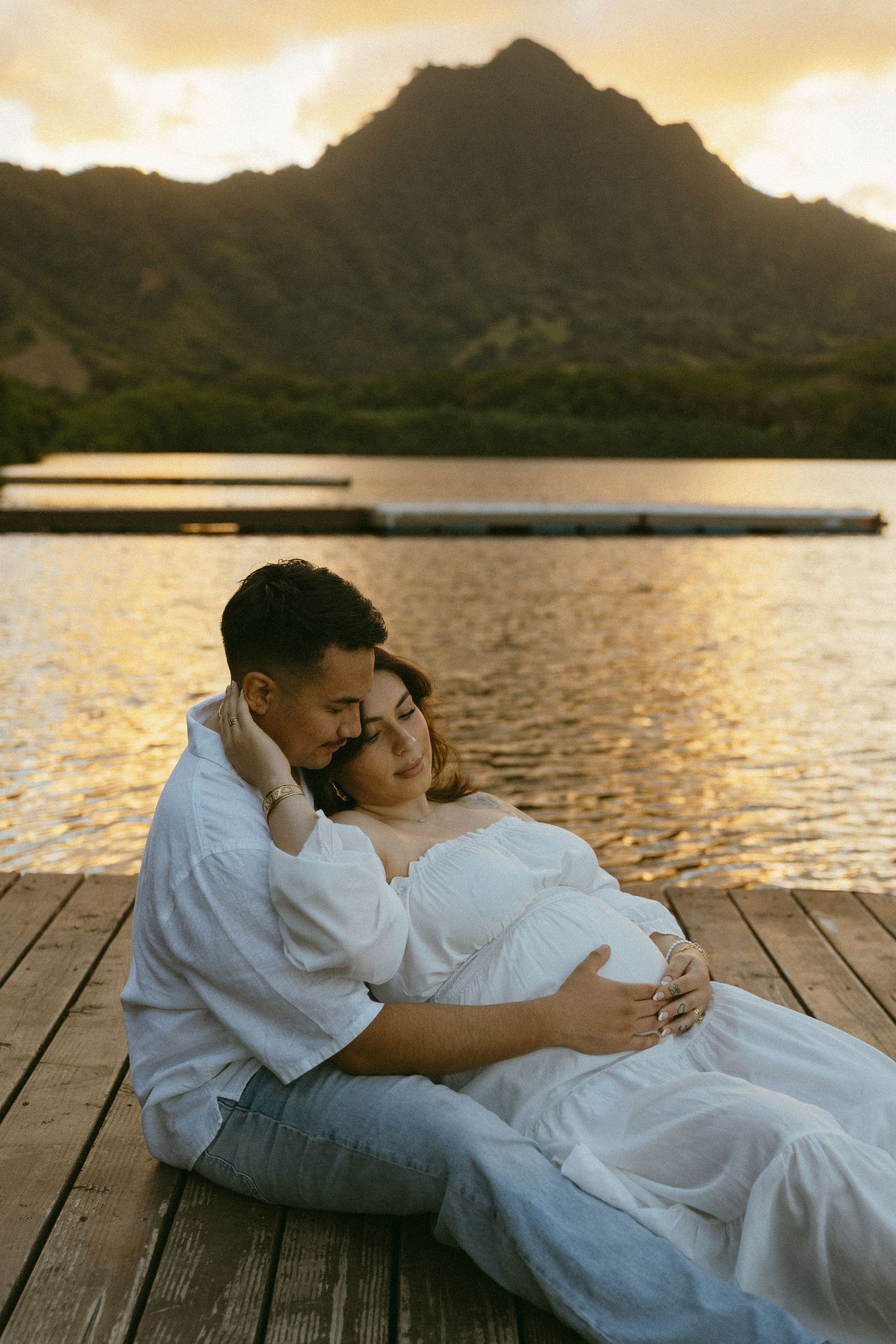 A couple in white clothing embraces on a wooden dock at sunset with mountains in the background.