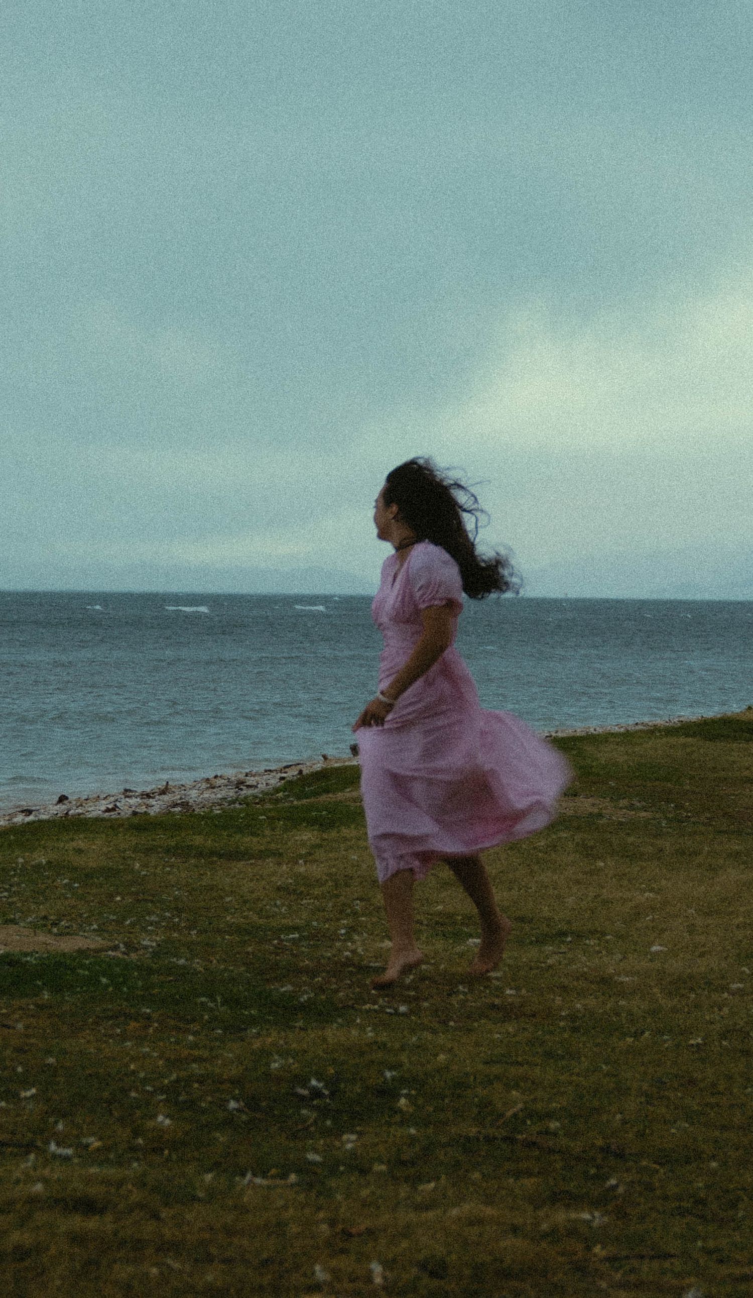 Figure in pink dress dances along coastal grassland with ocean in background.