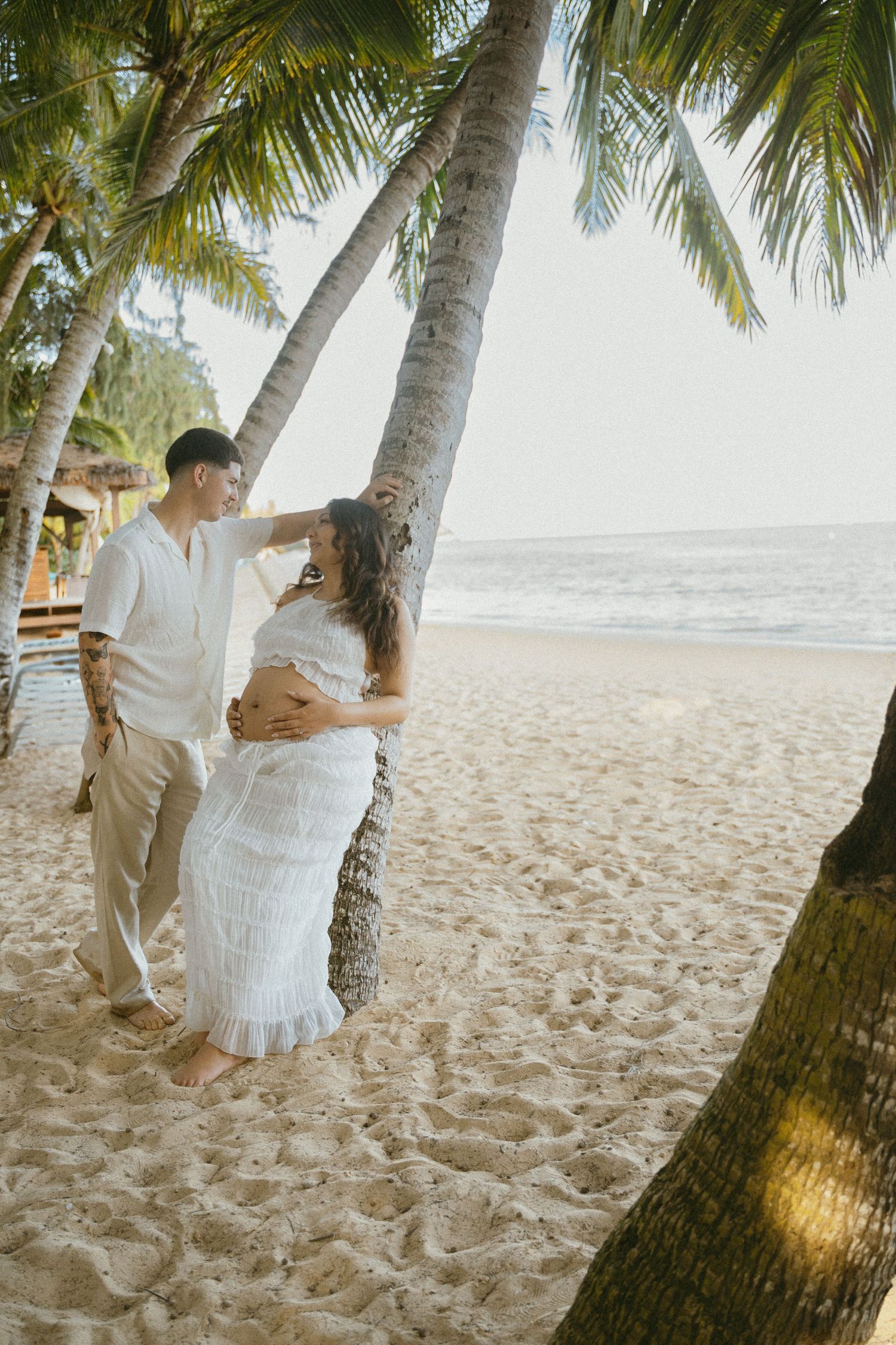 A couple poses under palm trees on a sandy tropical beach during a photoshoot.