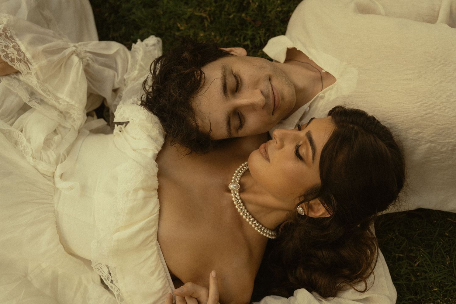 A couple in white clothing lies together on grass during a romantic photoshoot.