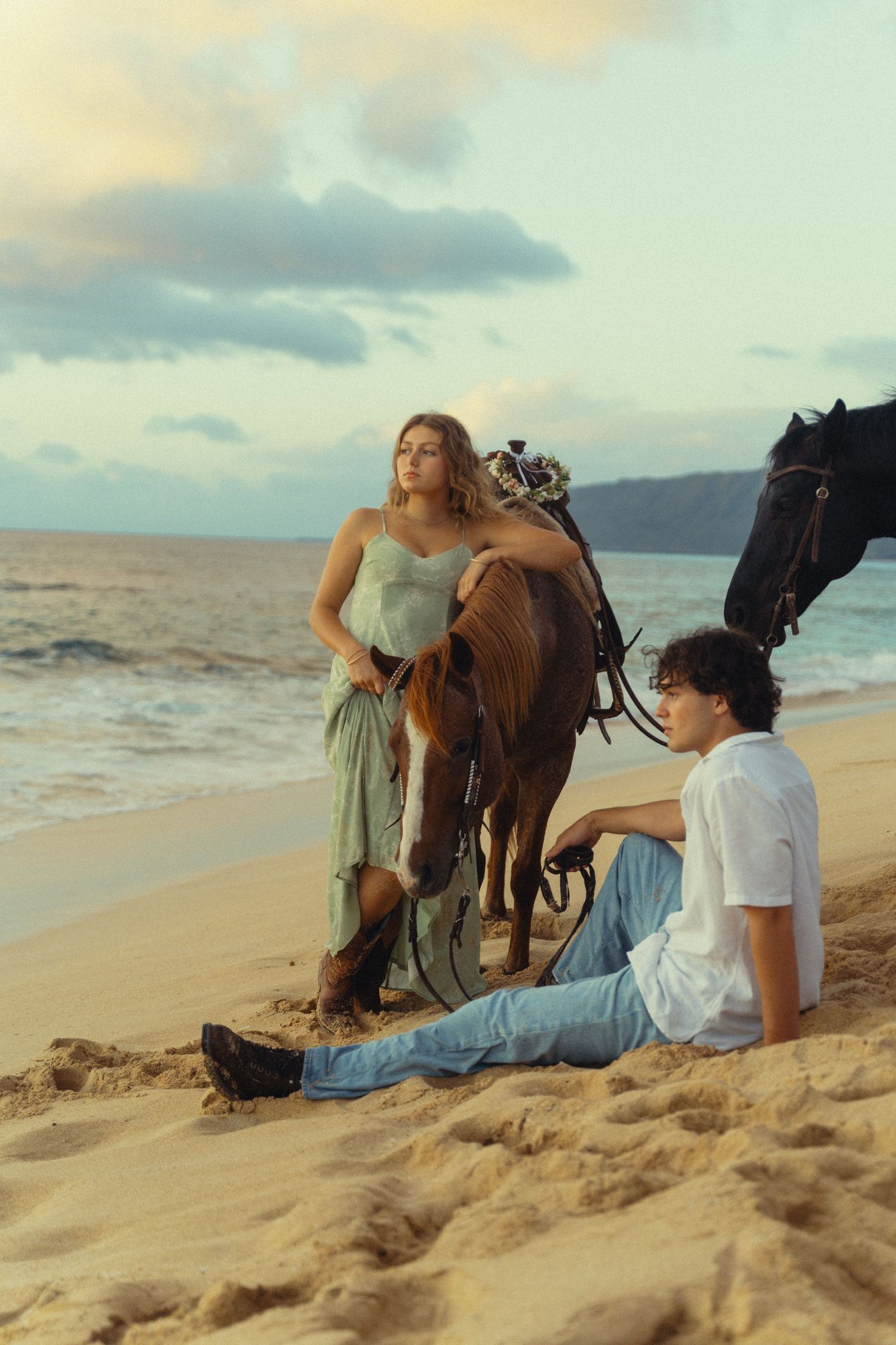 People relax on a sandy beach at sunset with horses and mountains in the background.