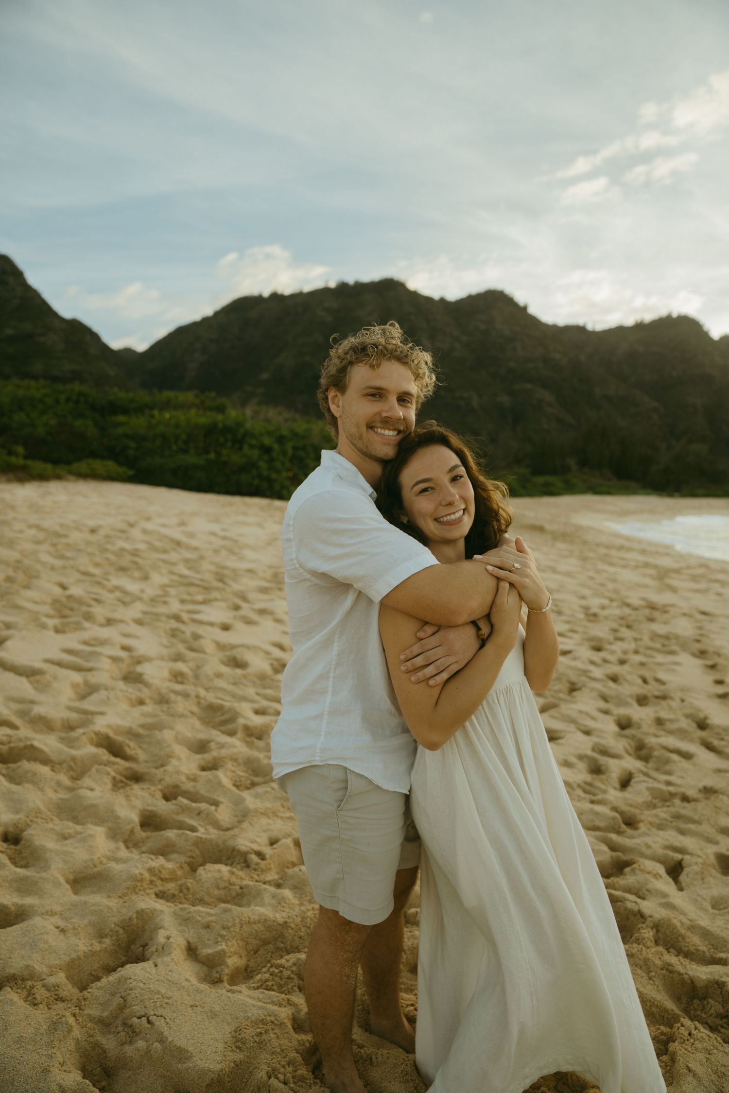 A joyful couple embracing on a sandy beach with dramatic mountain backdrop at sunset.
