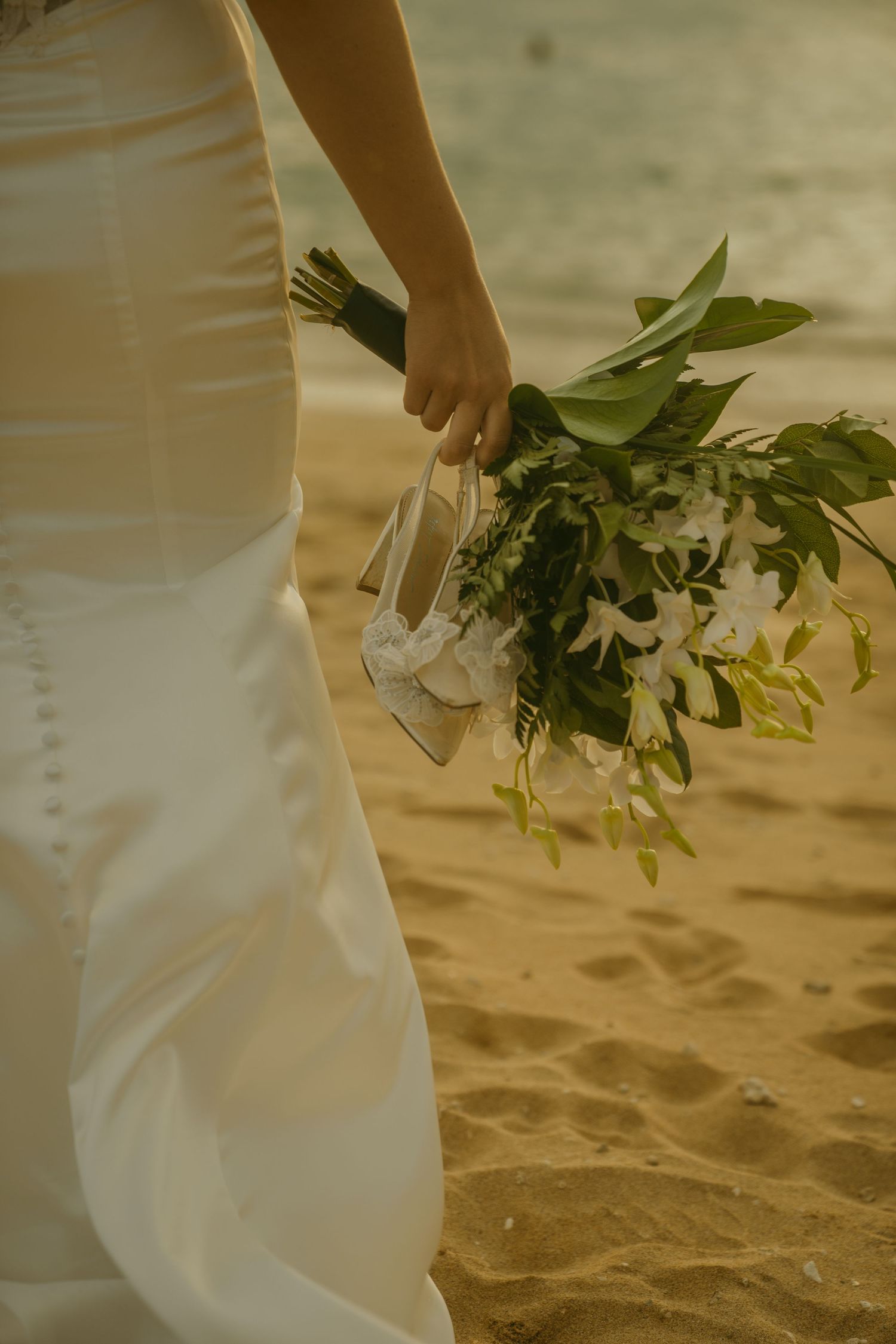 Close-up of white wedding dress and floral bouquet details on sandy beach.