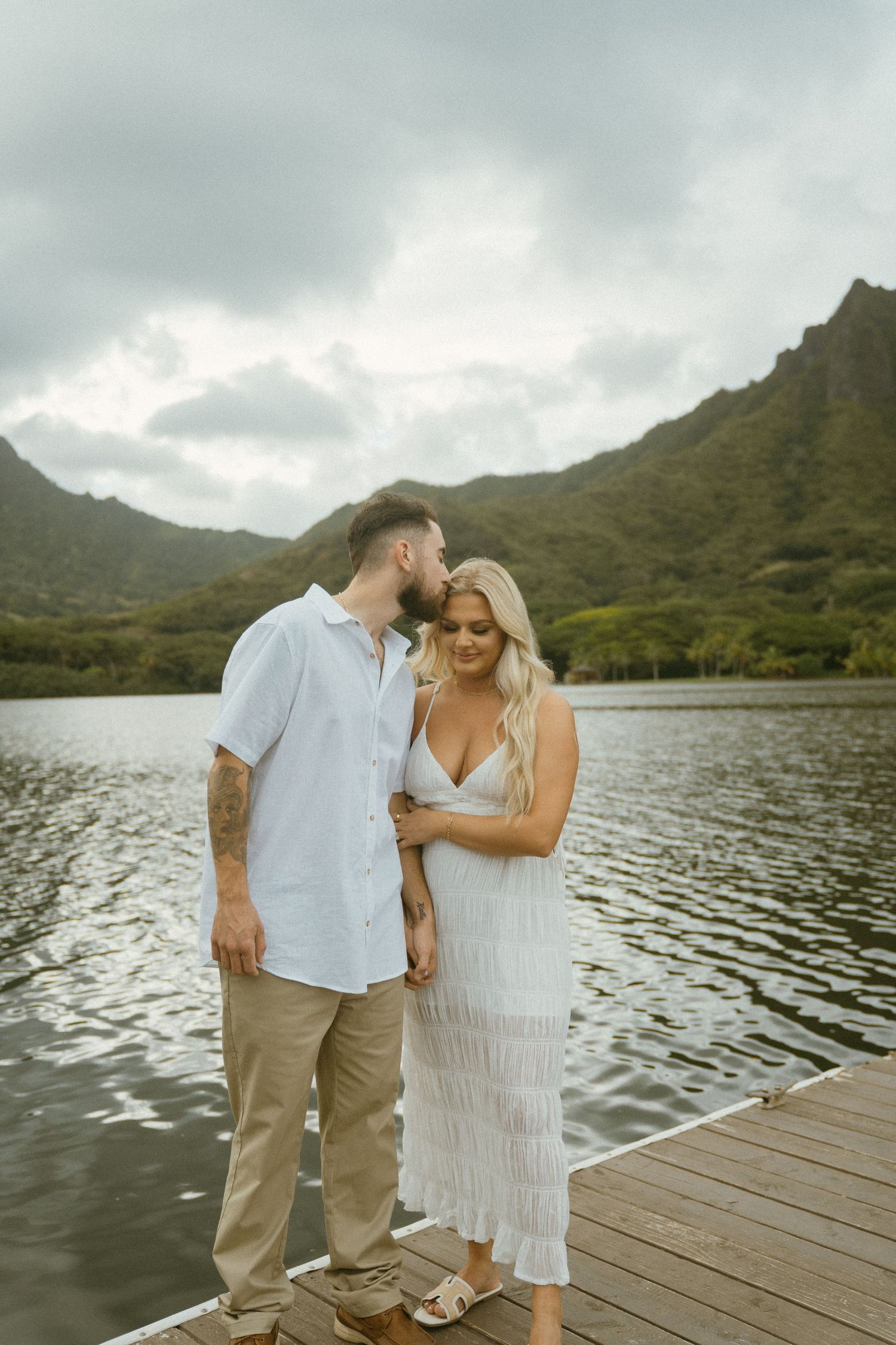 Couple in white clothing sharing tender moment on dock by mountain lake.