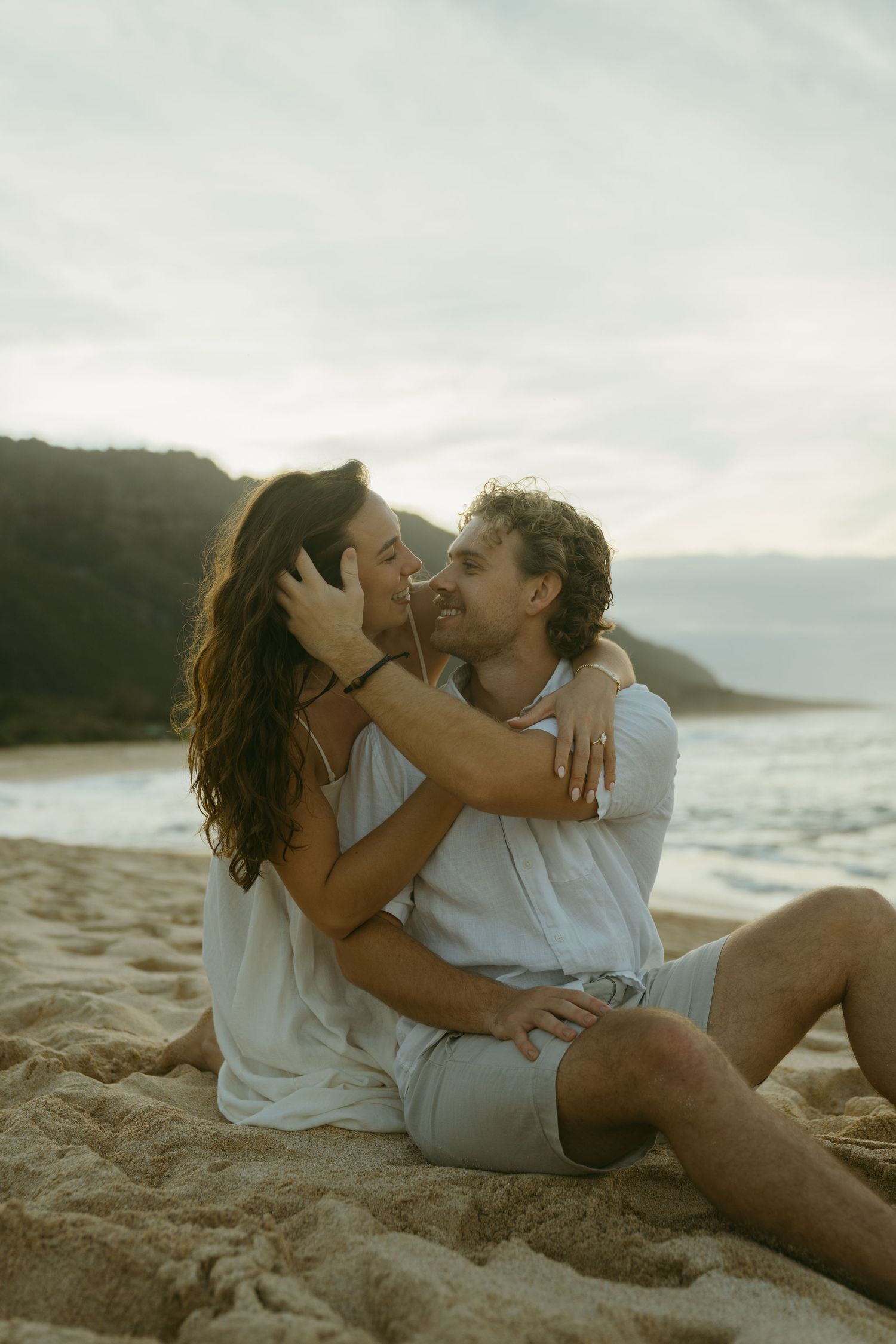 Couple in white clothing sharing romantic moments while sitting on sandy beach at sunset.