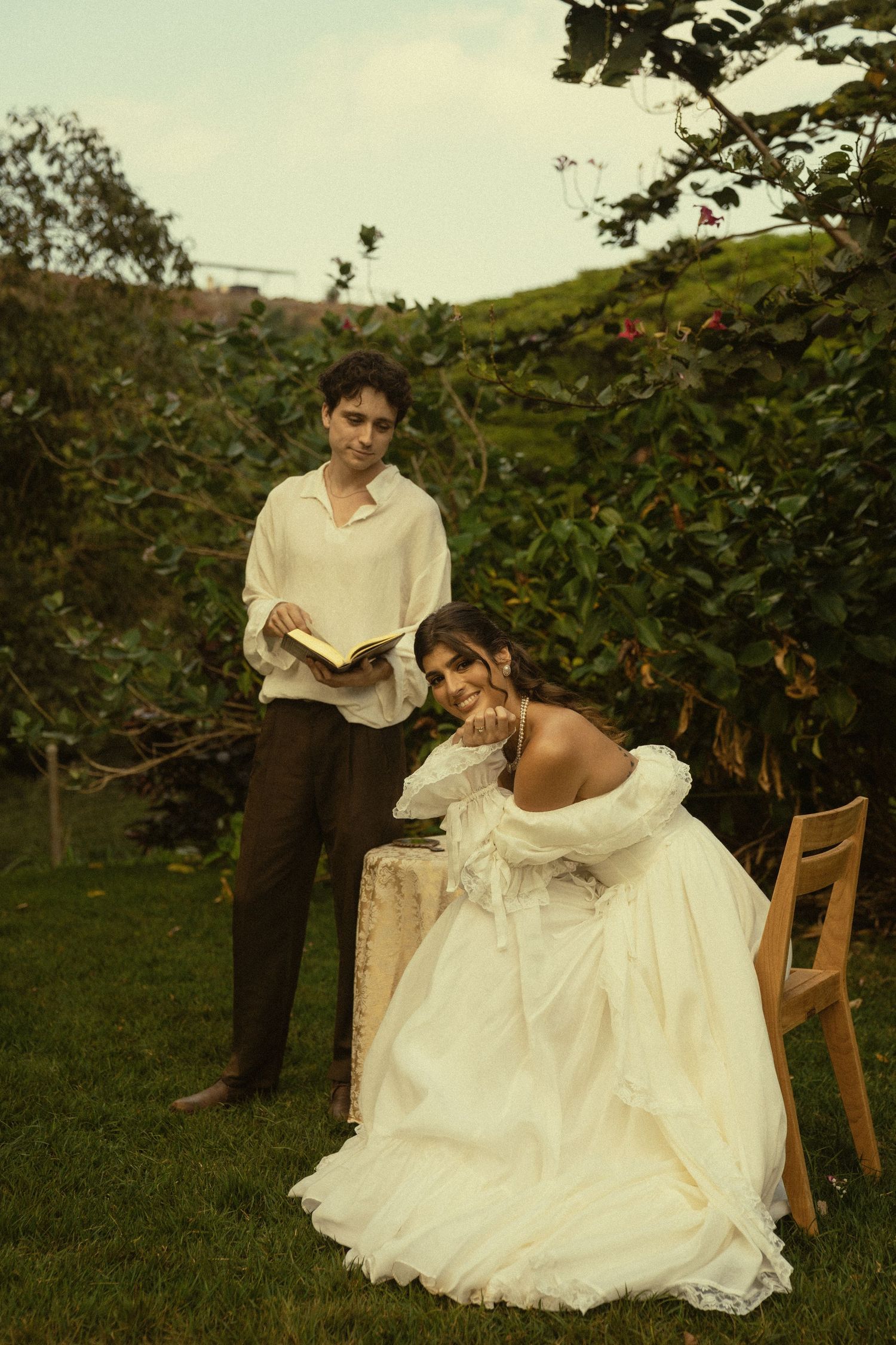A couple in vintage-style clothing poses for a romantic outdoor photoshoot in a garden setting.
