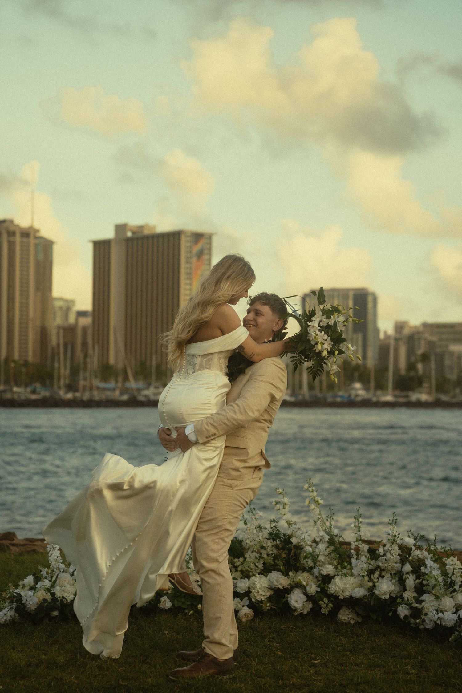 Newlyweds sharing romantic moment with city skyline across waterfront.
