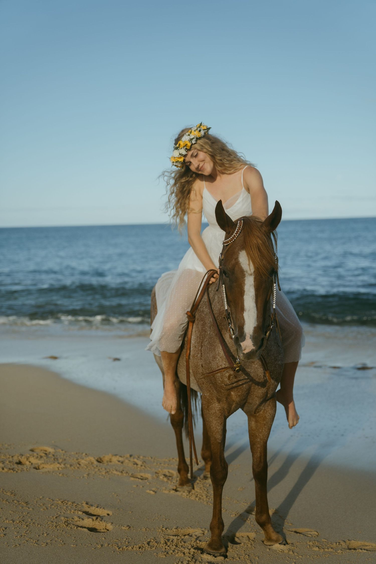 A woman in a white dress rides a brown horse along a scenic beach shoreline at sunset.