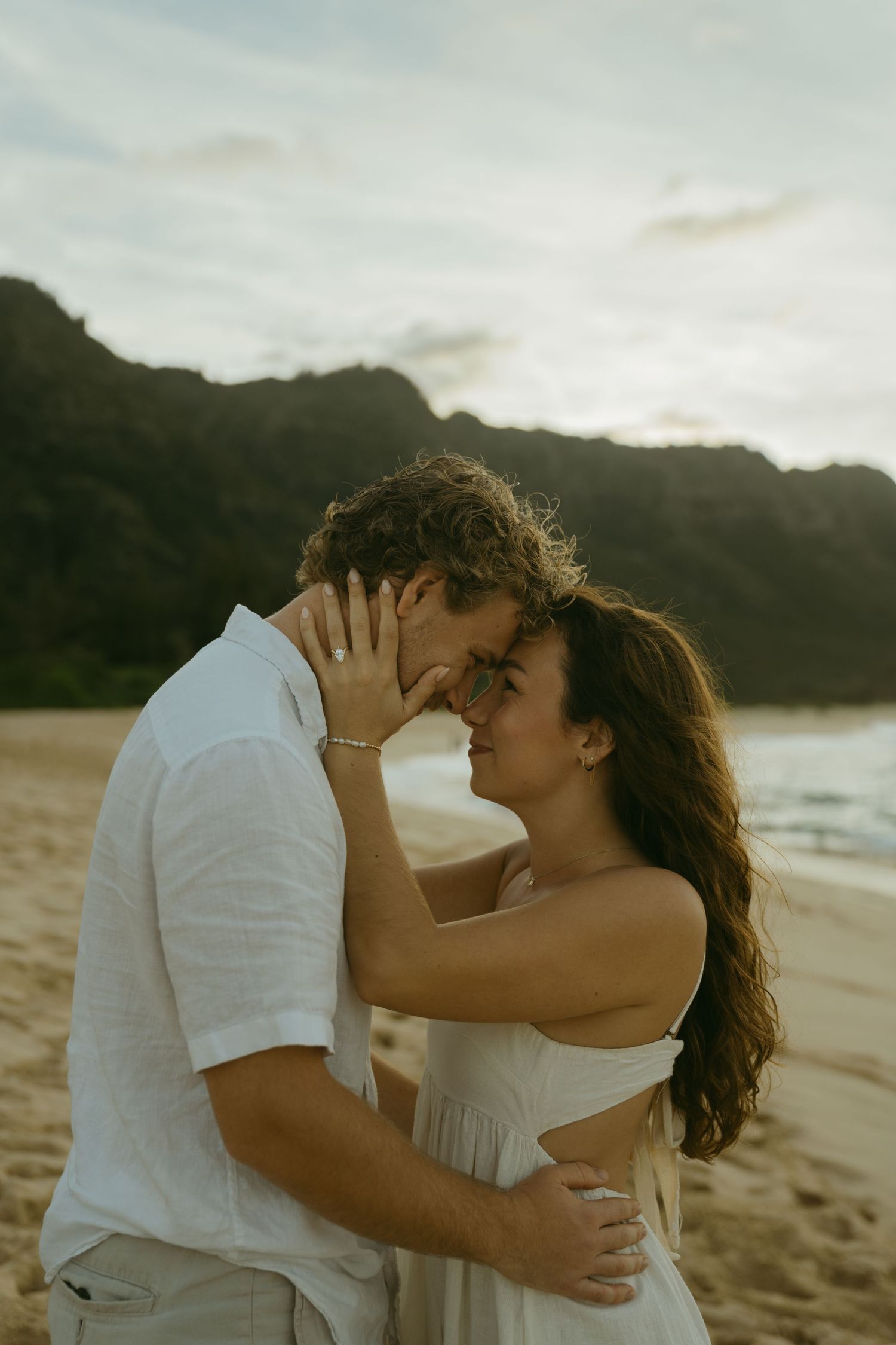 Couple embracing and kissing on a scenic beach with mountains in the background.