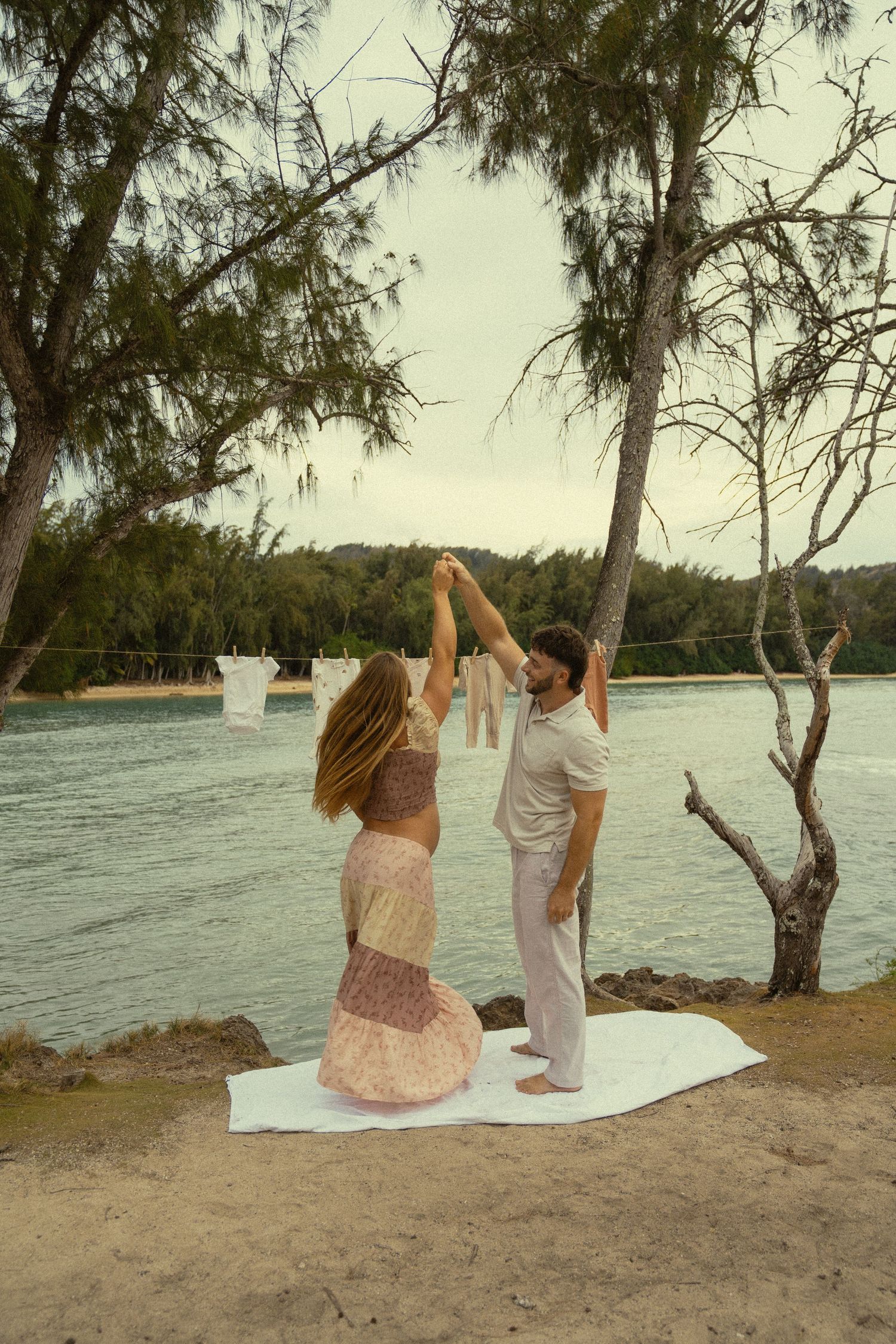 A couple sits on a white blanket by the beach at sunset with trees and water in the background.