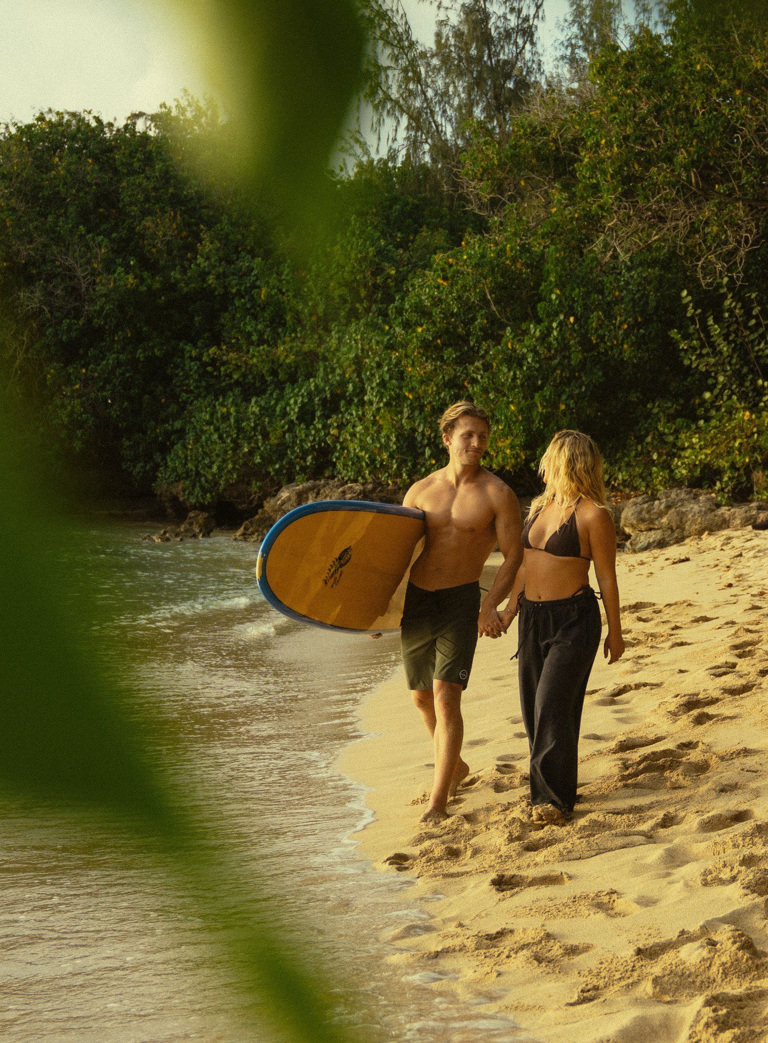 A couple walks along the shoreline carrying a surfboard with lush tropical vegetation in view.